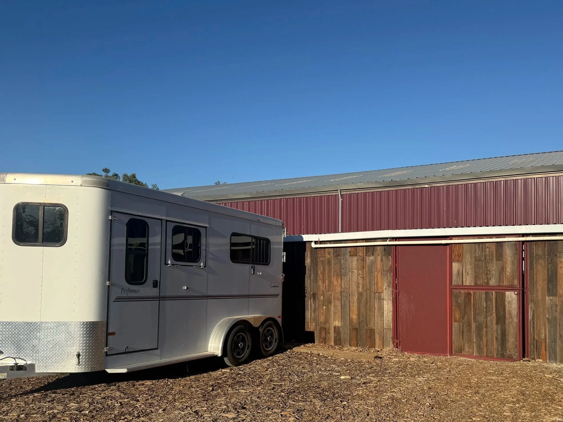 A white horse trailer parked next to a barn with red barn doors and a grey metal roof under a clear blue sky.