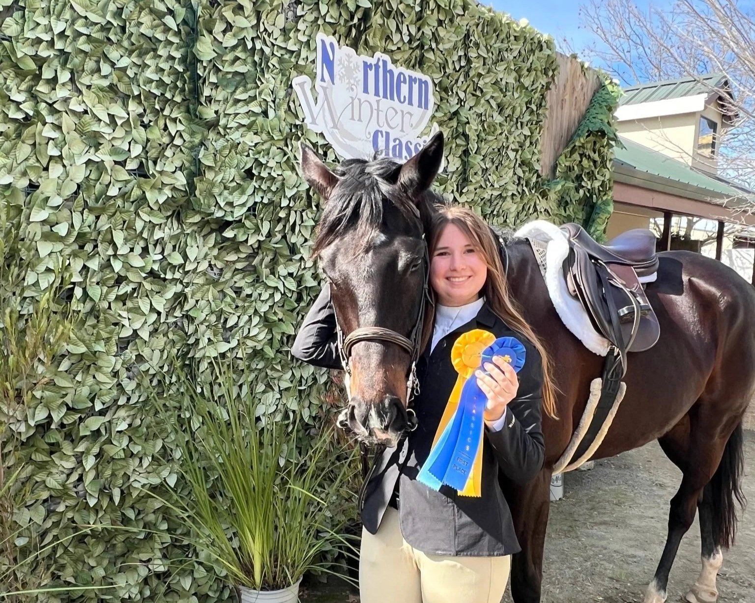 A young woman with long brown hair, smiling, standing next to a dark brown horse with a gray face. She is holding blue and yellow ribbons. They are outdoors in front of a green leafy hedge, with a sign that reads 'Northern Winter Classic' behind them.