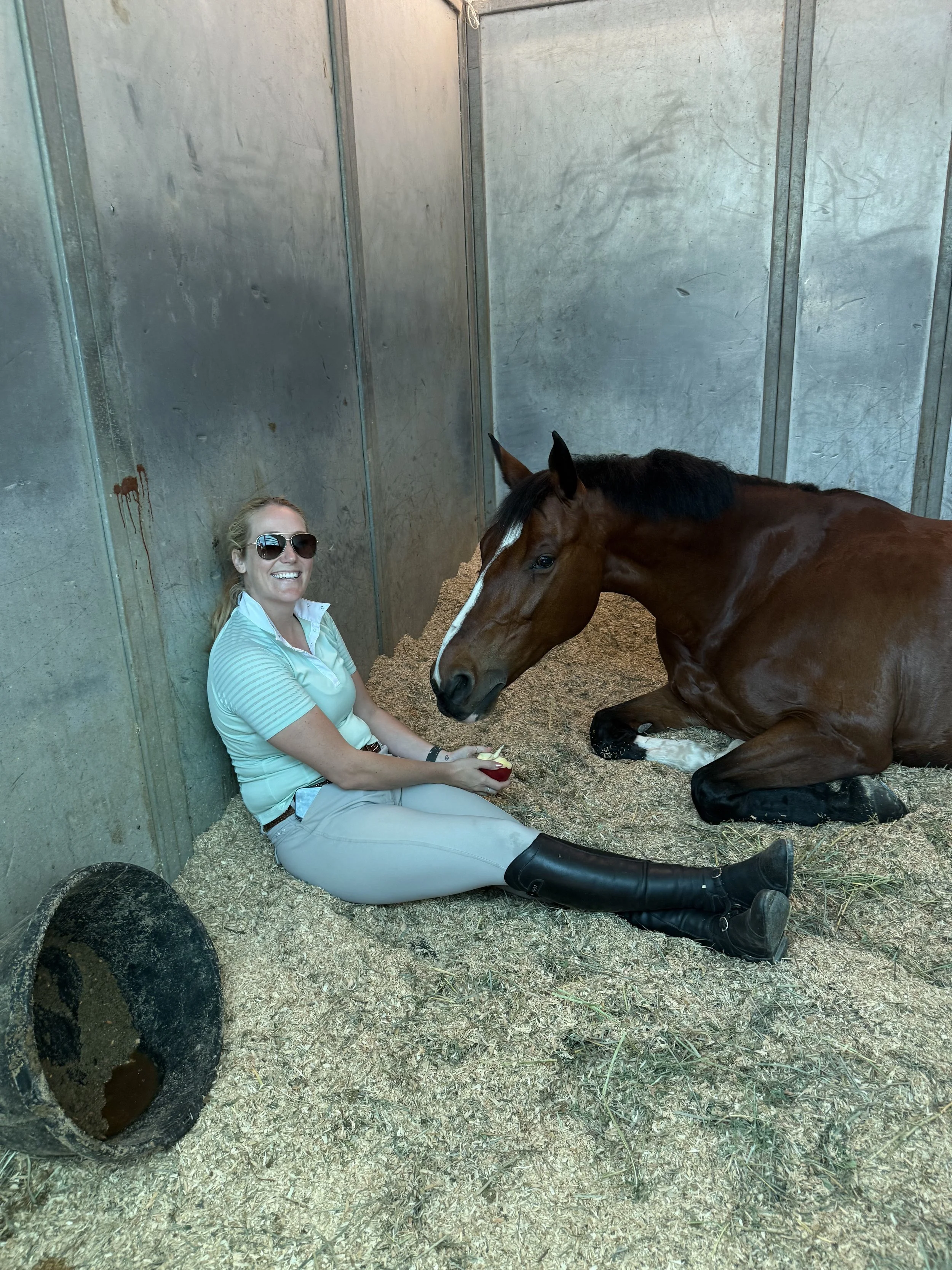 A woman sitting on the ground in a stable or barn, smiling and holding an apple, with a brown horse lying next to her. The woman is wearing sunglasses, a light-colored shirt, and riding boots. The stable has metal walls and a container of water on the ground.
