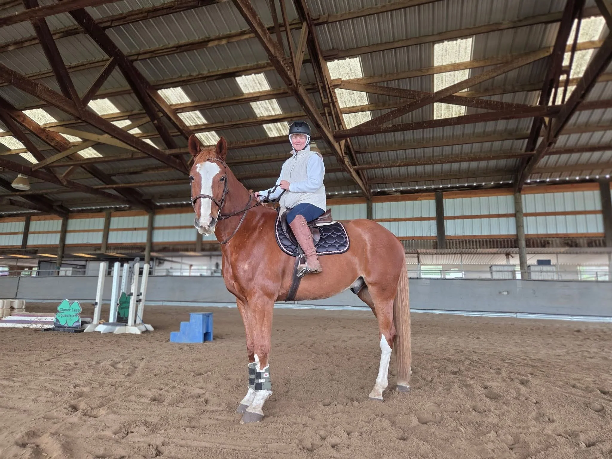 A woman wearing riding gear and a helmet is sitting on a chestnut horse inside an indoor riding arena. The arena has a high, metal roof with visible wooden beams and a dirt floor. There are some horse jumping obstacles in the background.