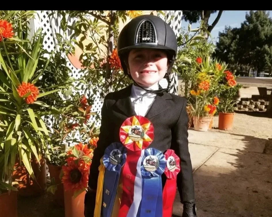 Young girl dressed in equestrian riding attire with ribbons on her chest, standing outdoors near potted plants and a white lattice fence, wearing a riding helmet, smiling.