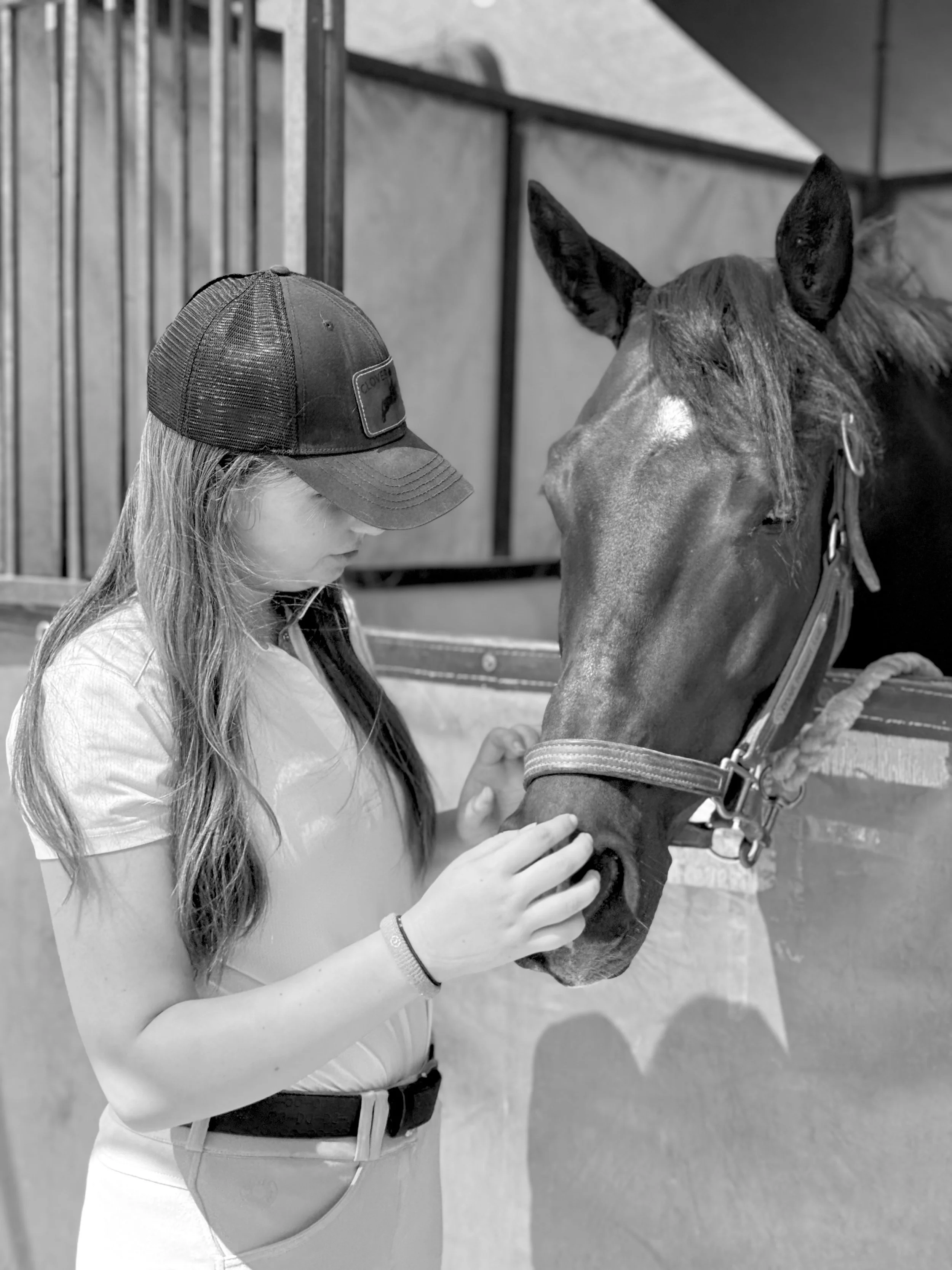 A young girl in a cap talks to a brown horse in a stable.