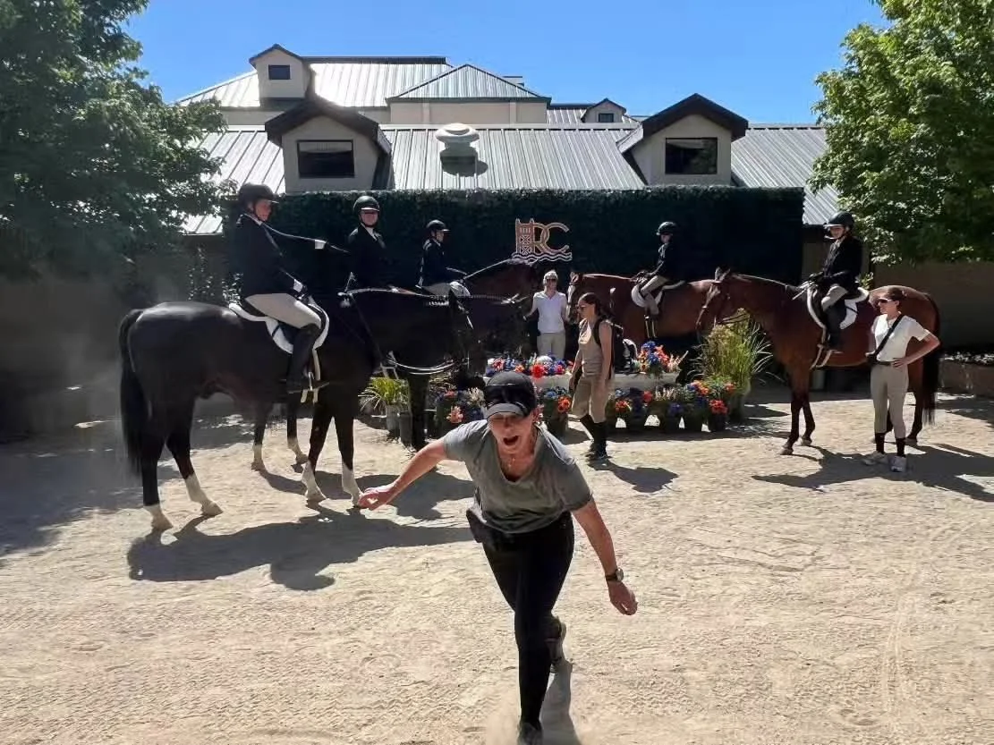 A woman in the foreground appears surprised or excited, running towards the camera, while five riders on horses are positioned behind her with a decorative setup of flowers and plants, near a building with a metal roof and trees on either side.