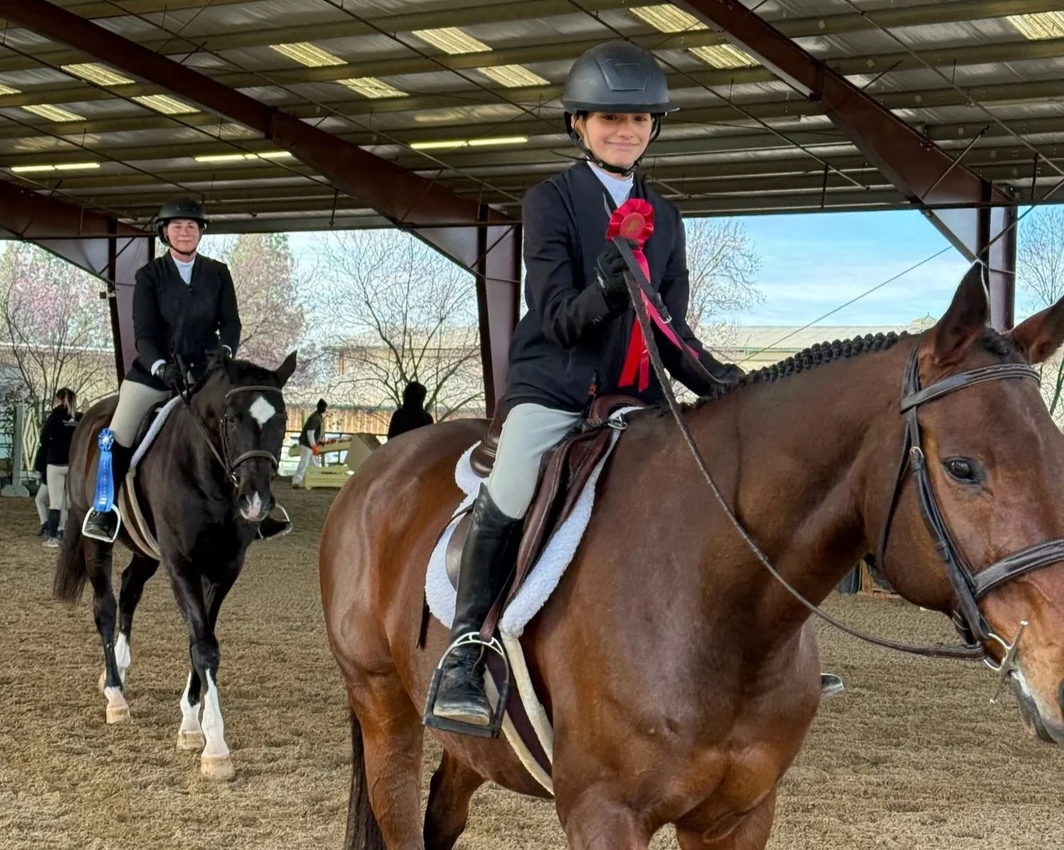 Two young girls in riding gear and helmets sitting on horses inside an indoor arena, with medals and ribbons indicating a competitive event.