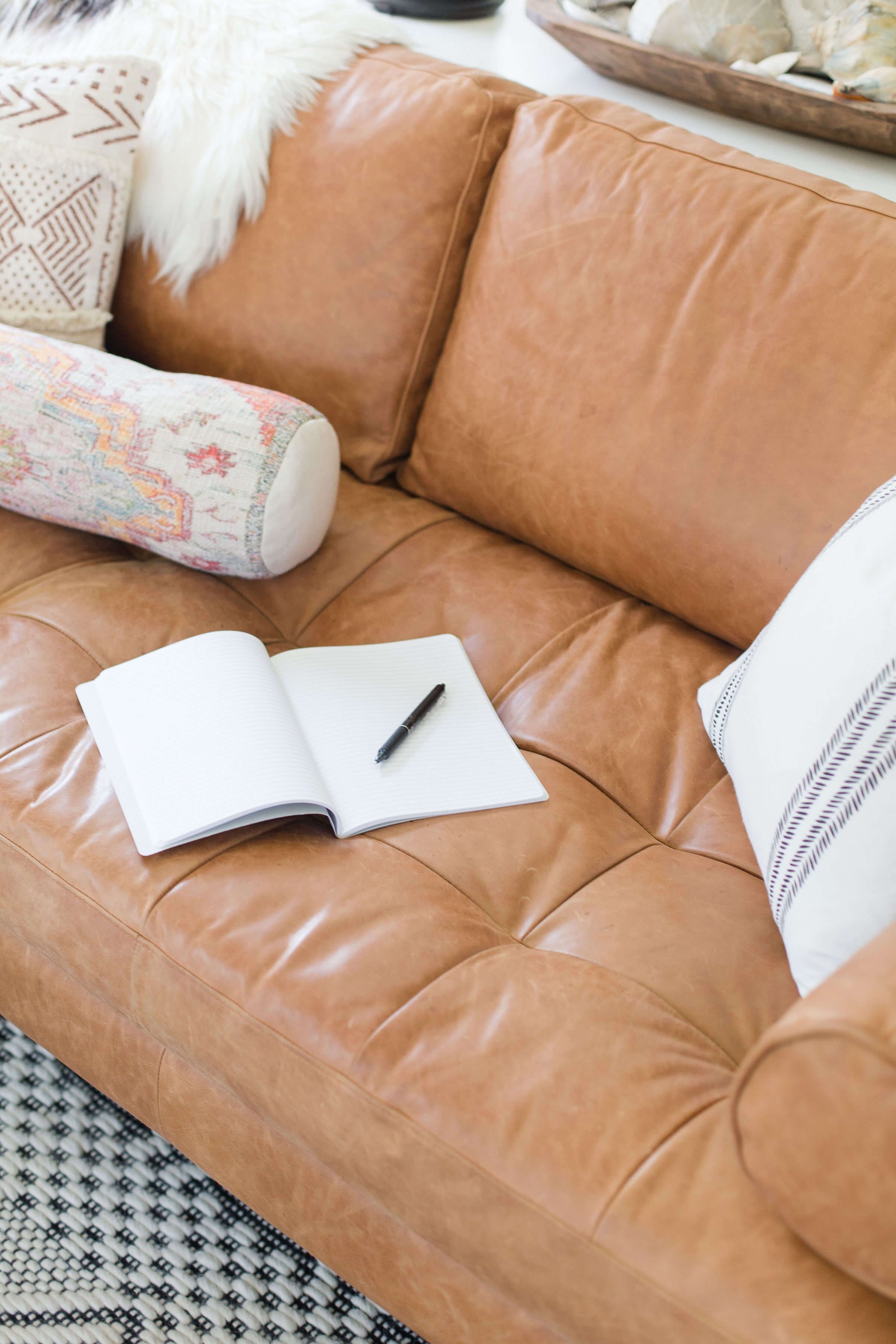 light brown leather couch with an open notebook and pen laying on it. Pillows and blankets in the background