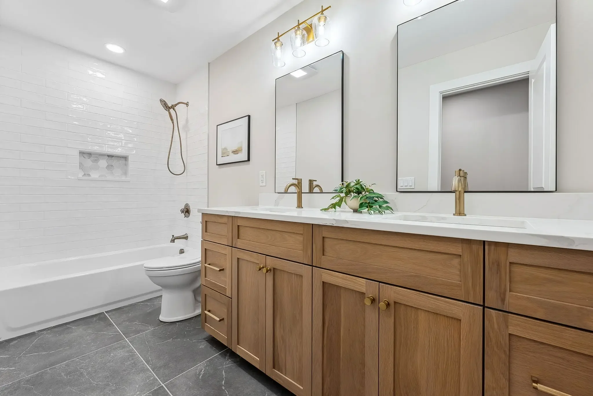Modern bathroom with a wooden vanity, large mirror, and white countertops, featuring a plant, brass fixtures, a white bathtub, and a shower area with white subway tiles.