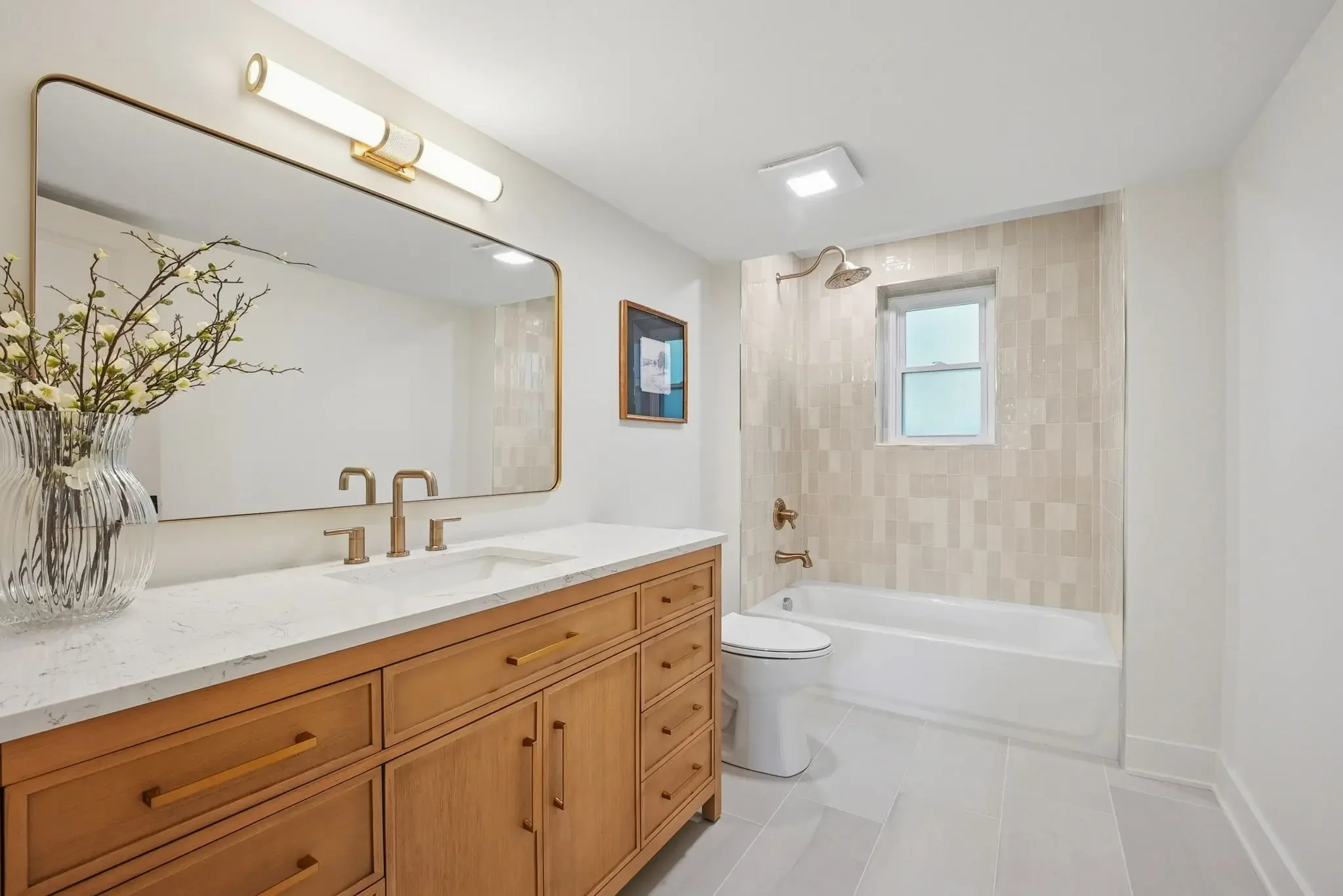 A bathroom with a large vanity mirror, a white countertop with a vase of flowers, a wooden cabinet, a sink with gold fixtures, a framed picture on the wall, a shower area with beige tiles, a window, a showerhead, a bathtub, and a toilet.