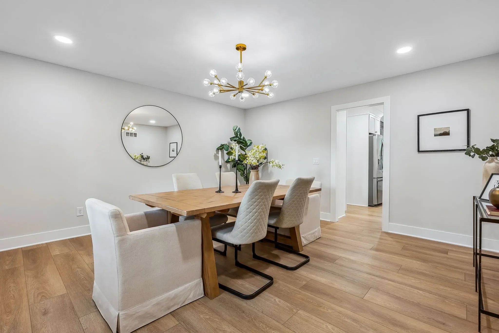Dining room with a wooden table and six upholstered chairs, a modern chandelier, a large round mirror on the wall, and decorative plants.