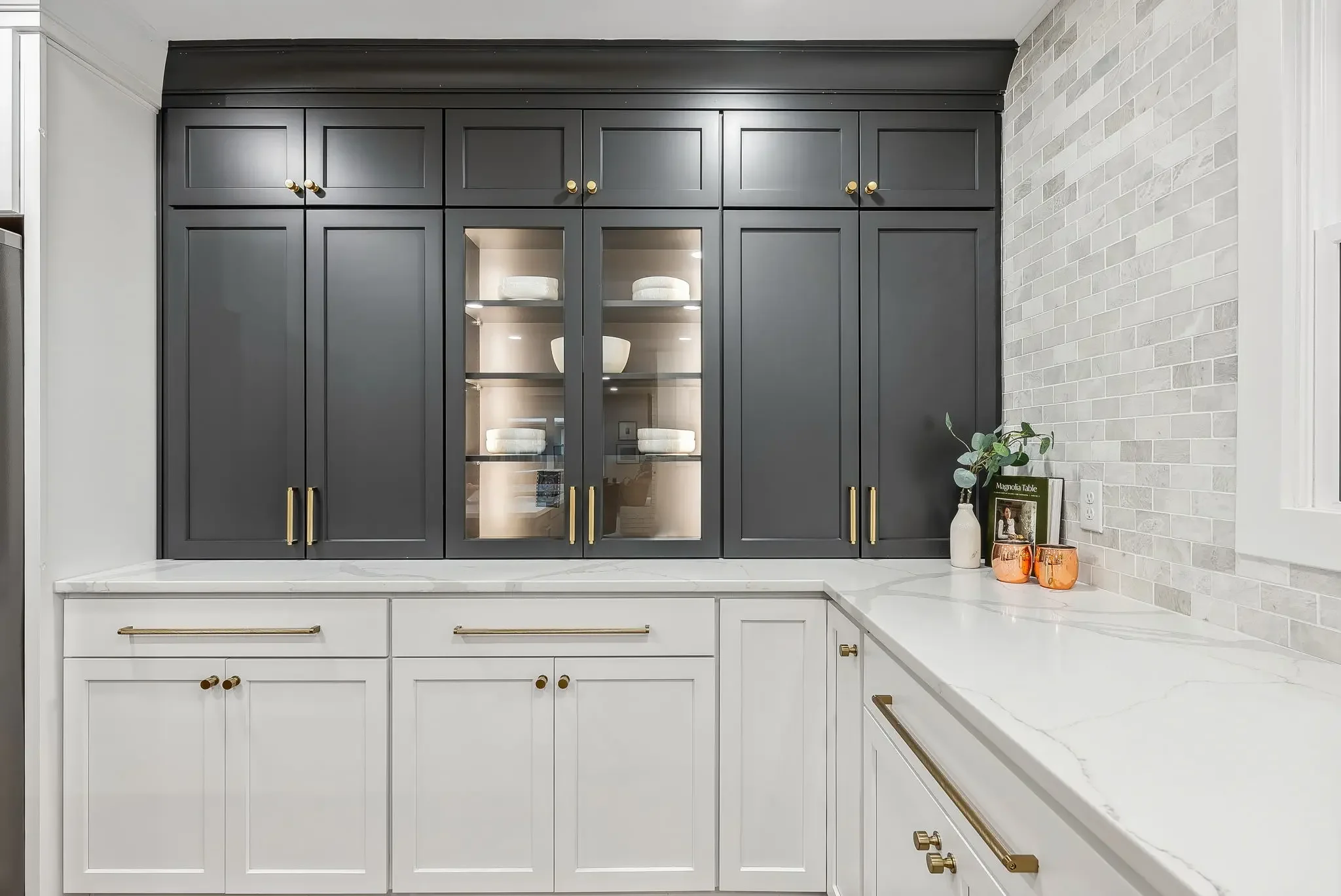 Kitchen with dark gray upper cabinets with glass doors displaying white dishes and gold handles, white lower cabinets with gold handles, white marble countertops, a white brick backsplash, and decorative vases and books on the counter.