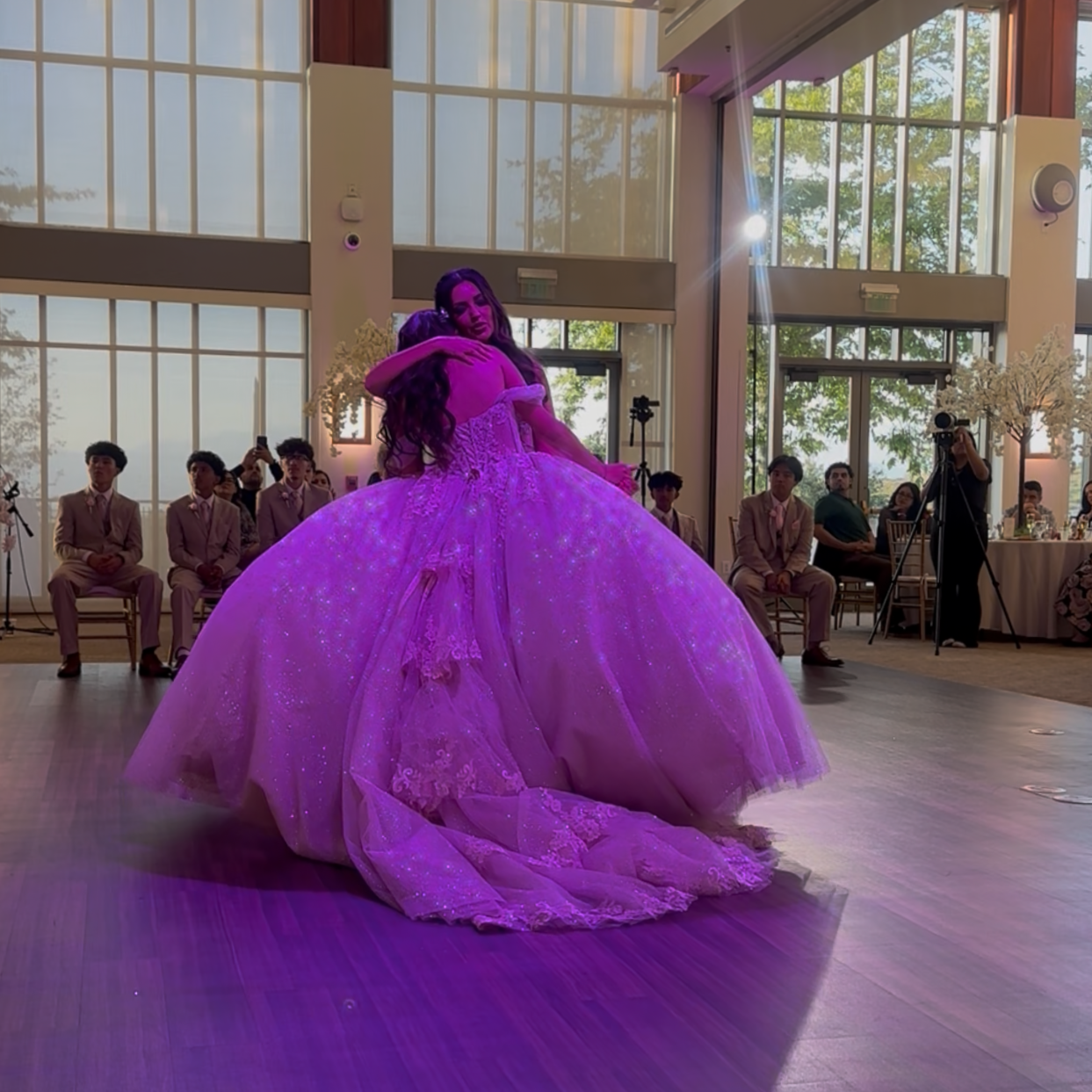 A bride and groom embrace during their wedding dance in a bright reception hall with large windows, seated guests, and photographers capturing the moment.