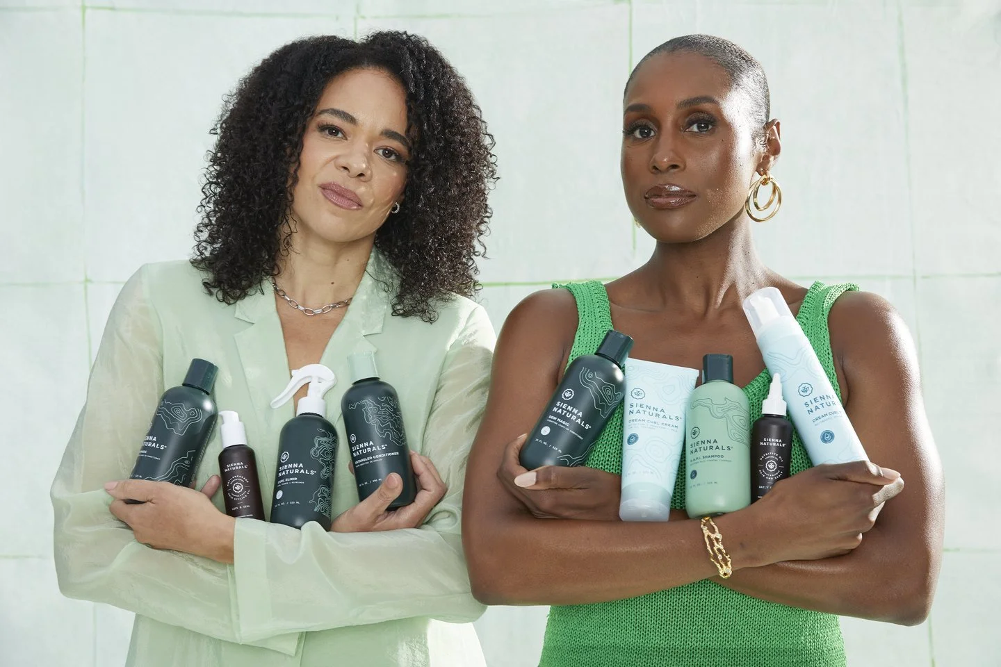 Two women standing against a light green tiled wall, holding various personal care products from Sienna Naturals, with confident expressions.