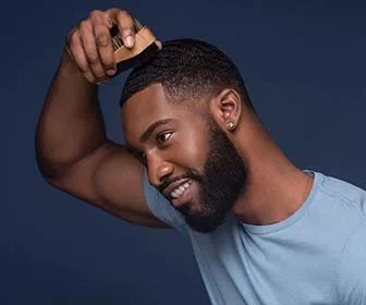 A smiling man with a beard and short hair, wearing a light blue shirt, touching his head with his hand against a dark blue background.