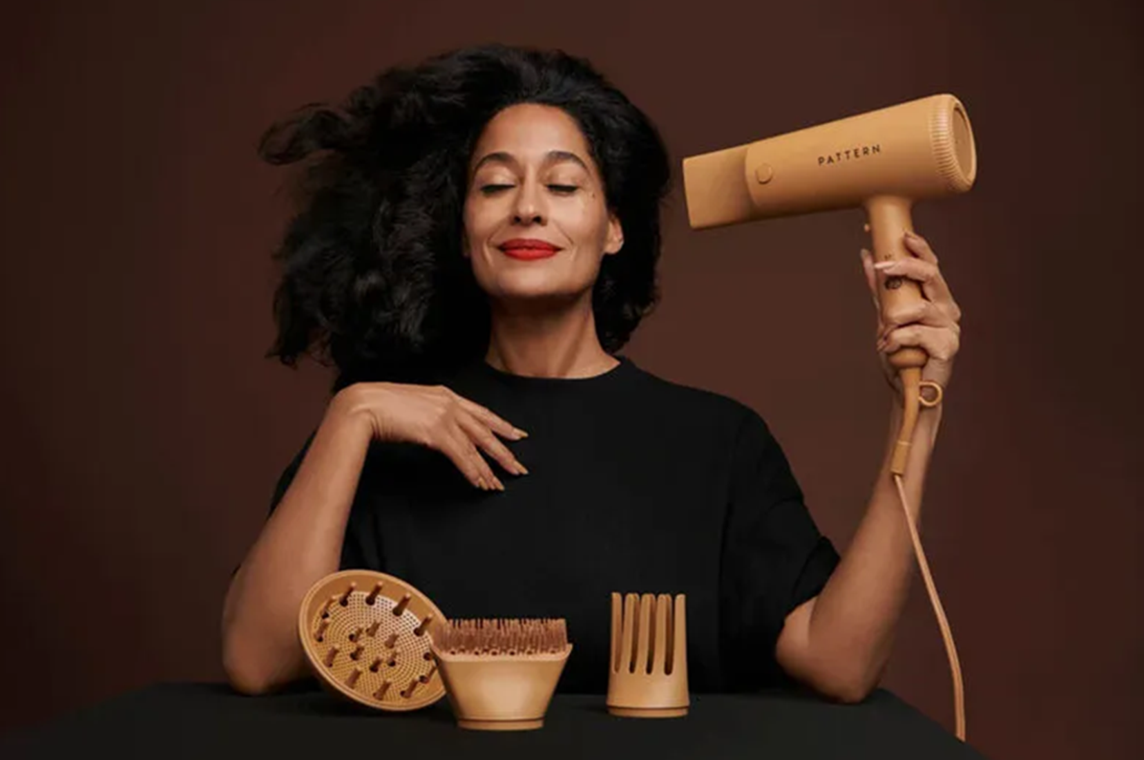 A woman with black curly hair and red lipstick smiling with her eyes closed, holding a beige hair dryer labeled 'Pattern' in her right hand, with various wooden hair styling tools on a black surface in front of her, against a brown background.