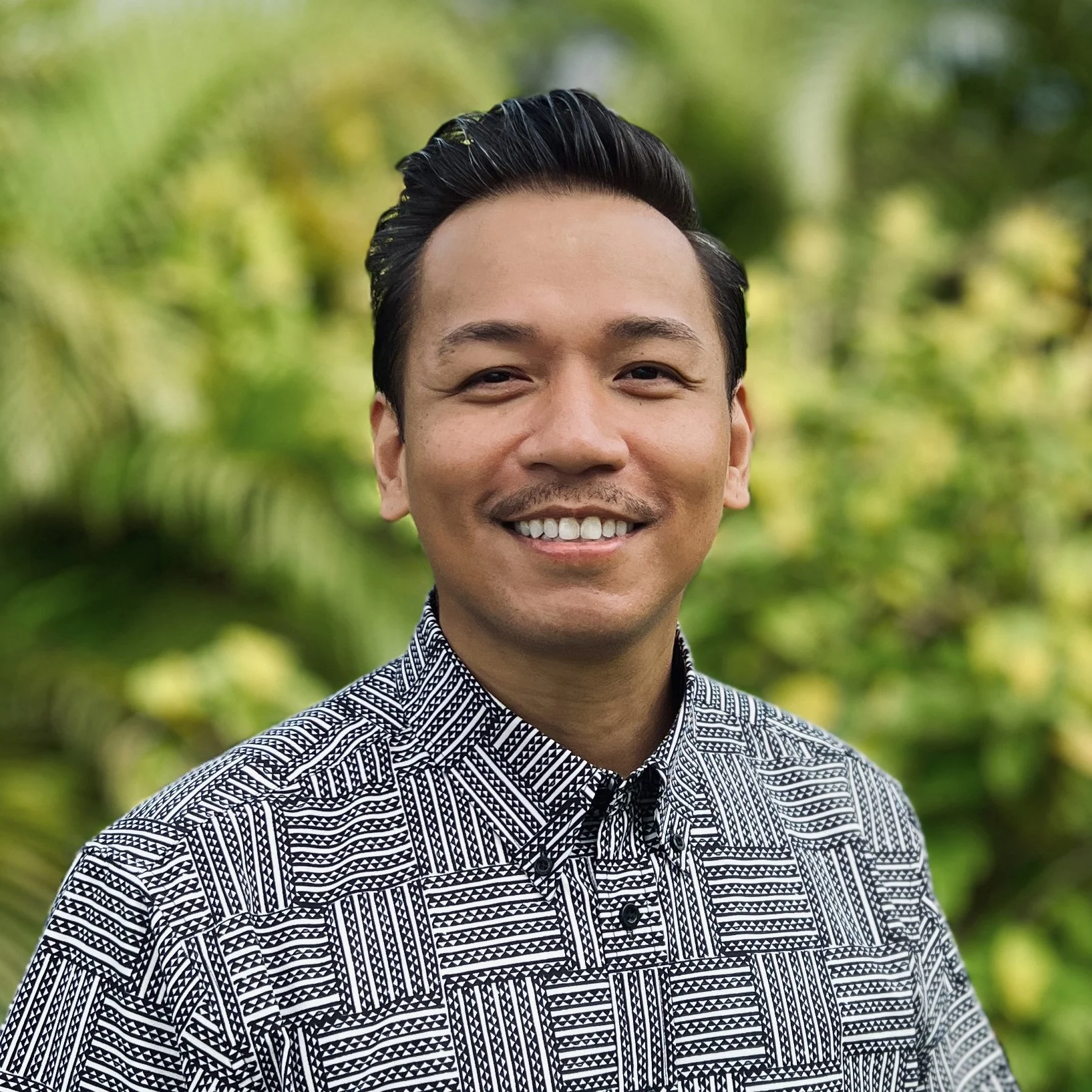 A smiling man with dark, slicked-back hair wearing a patterned black and white shirt standing outdoors with a lush green background.