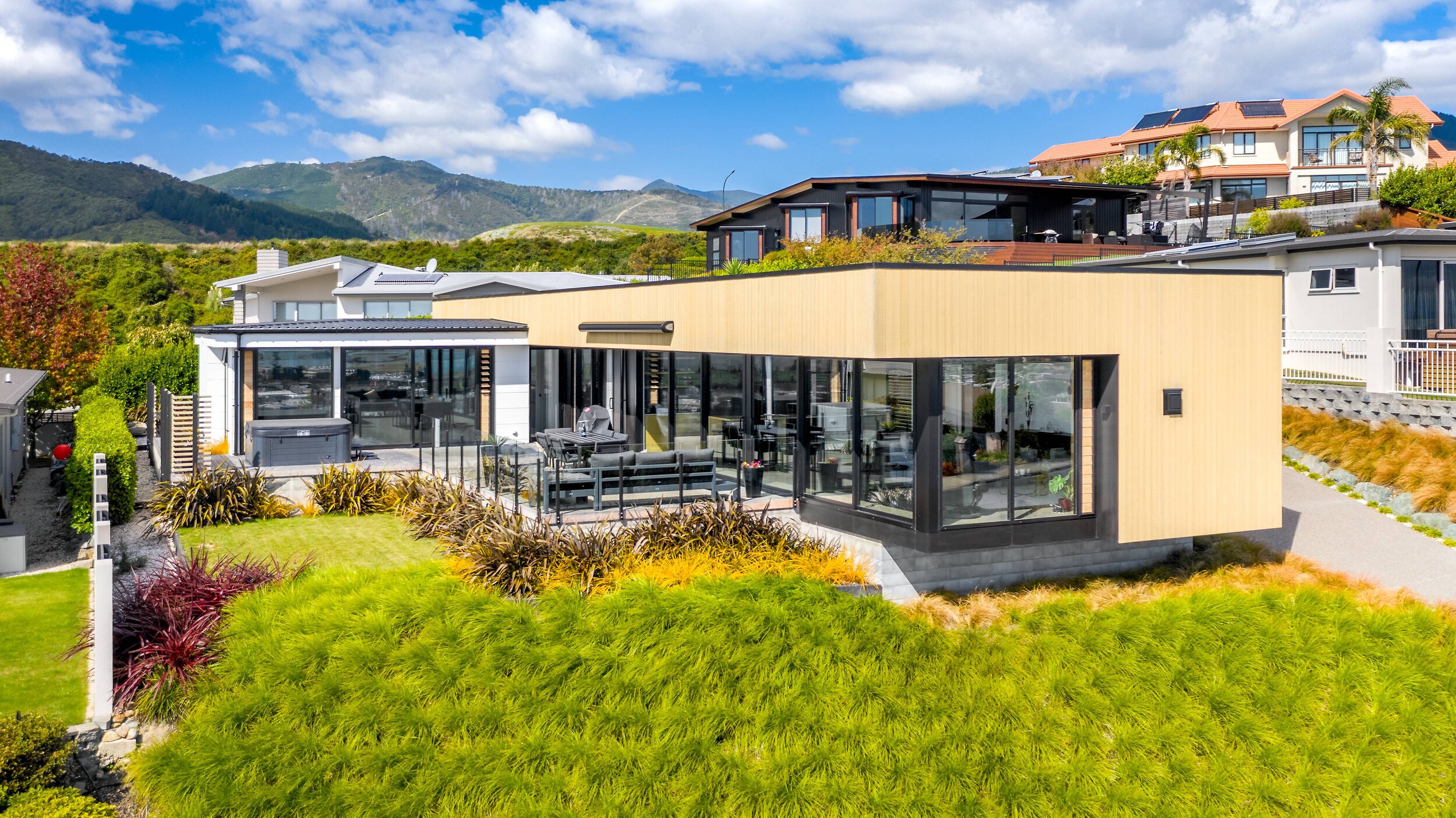 Modern house with large glass windows and outdoor patio surrounded by landscaped garden, with neighboring houses on a hillside under a partly cloudy sky.