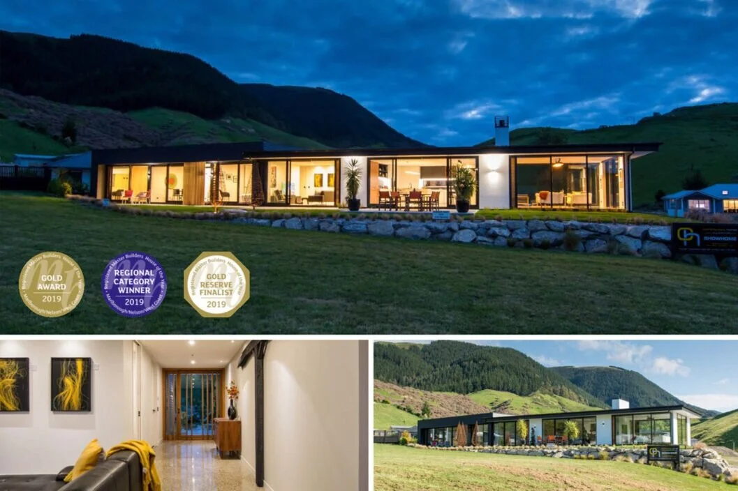 Modern house with large glass windows, situated on green grass with hills in the background. The house is illuminated from within, showing interior furnishings. The house has received awards for architecture in 2019.