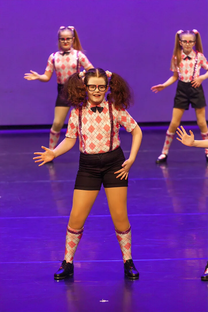 Young girl with glasses and pigtails performing a dance on stage with two other girls in similar costumes in the background.