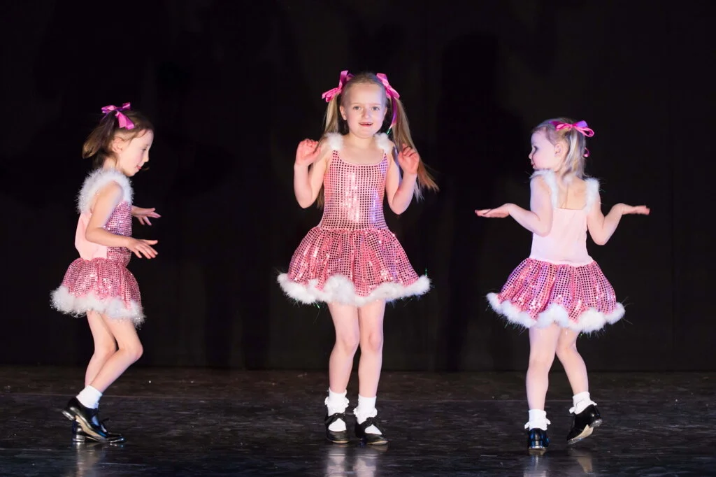 Three young girls in pink and white costumes dancing on stage.