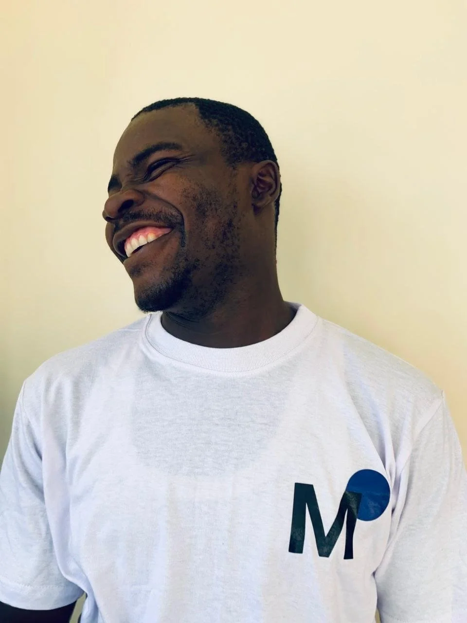 Ein lachender Mann mit dunkler Hautfarbe trägt ein weißes T-Shirt mit einem Logo, er hat kurze Haare und steht vor einem hellen Hintergrund.