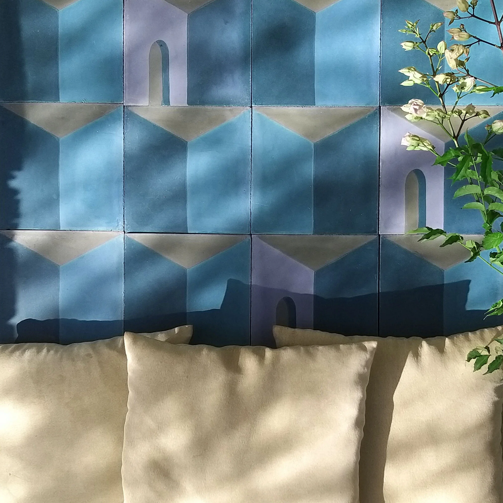 A wall with blue, purple, and beige tiles arranged in a geometric pattern resembling arches and cubes, with a beige sofa in front and a leafy plant partly visible on the right side.