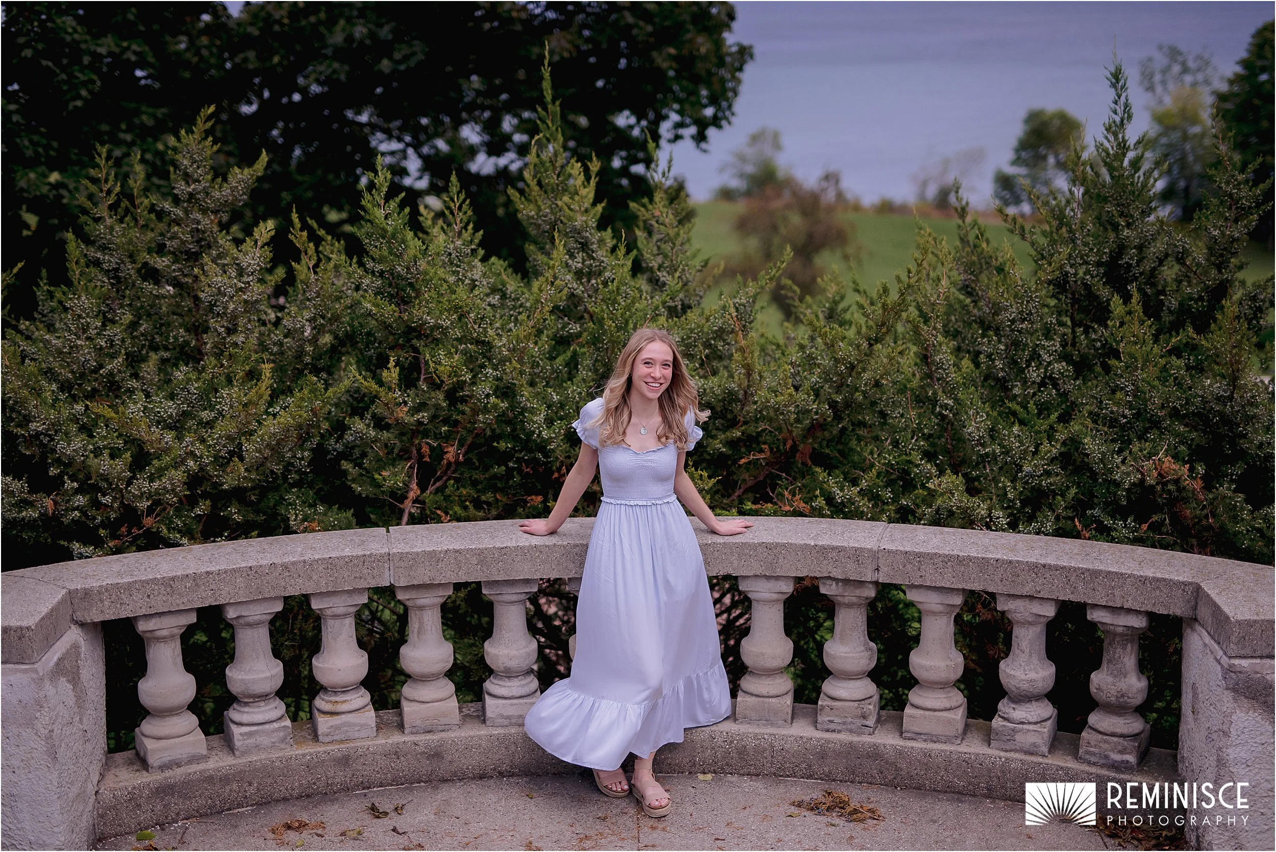 09-senior-leaning-stone-pillar-railing-lake-green-bushes-background.JPG