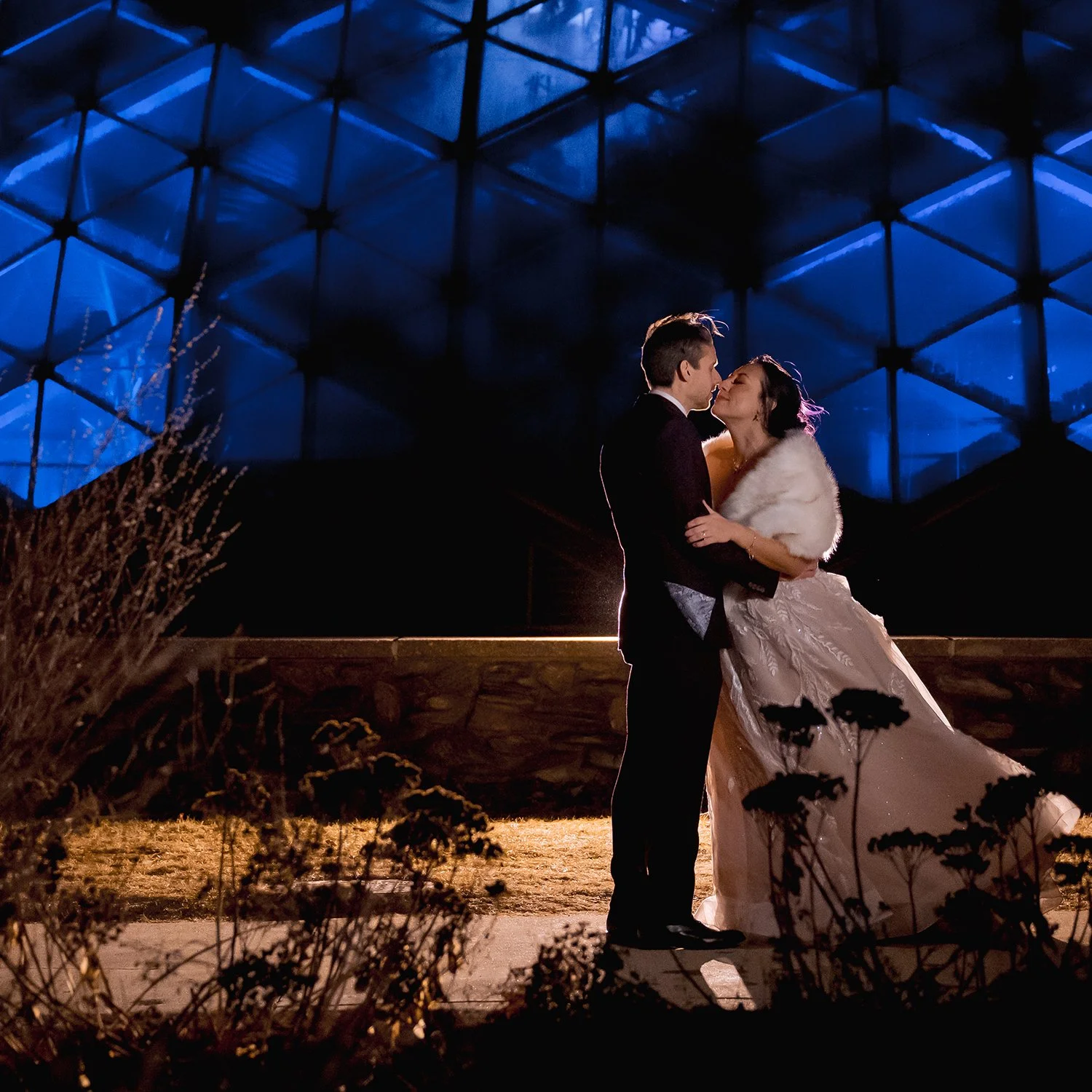 Bride and groom sharing a kiss during an artistic nighttime wedding portrait in Milwaukee with dramatic blue architectural lighting at the mitchell park domes