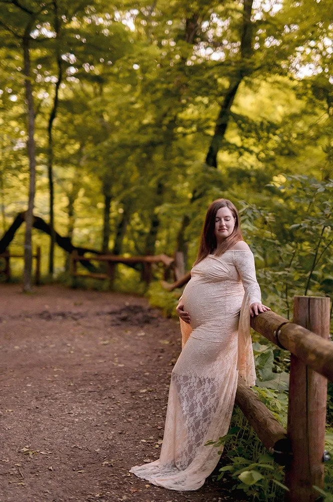 Expecting mother in a lace gown leaning against a wooden fence along a wooded trail in Milwaukee during a natural maternity photography session at Grant Park's Seven Bridges