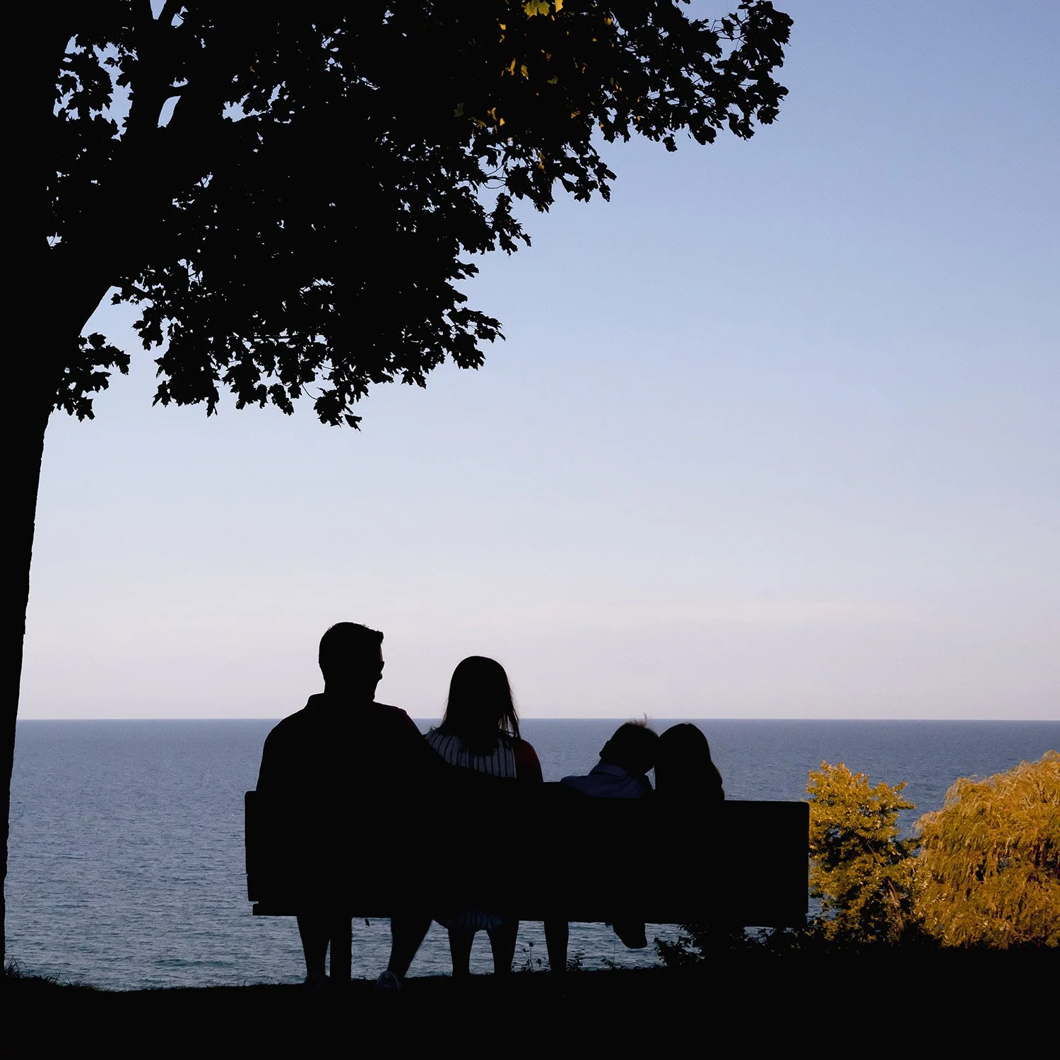 Silhouette of a family of four sitting together on a bench overlooking Lake Michigan during a Milwaukee family photography session