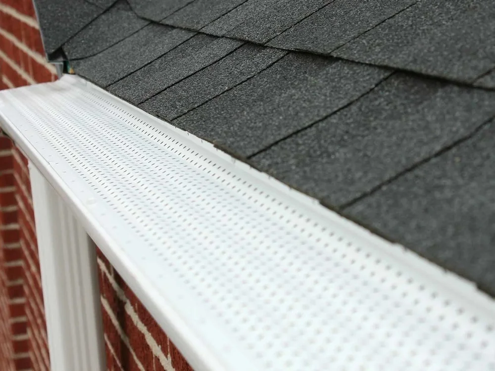 White perforated gutter guard installed along a shingle roof edge on a brick home.