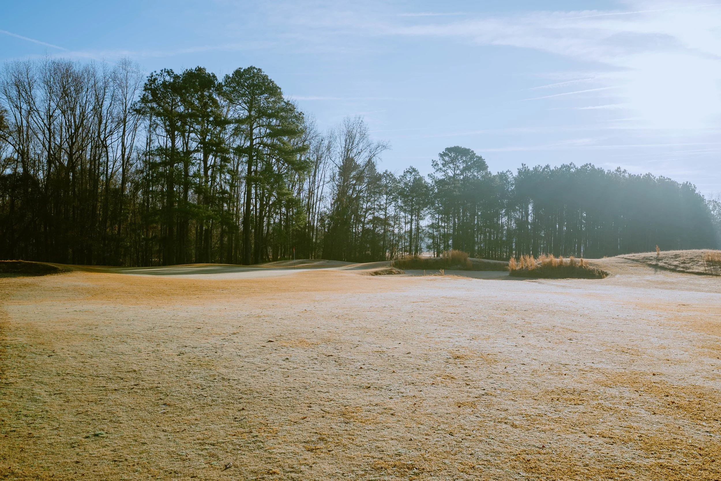 A golf course with sand traps and a distant tree line under a clear blue sky, with the sun shining brightly.