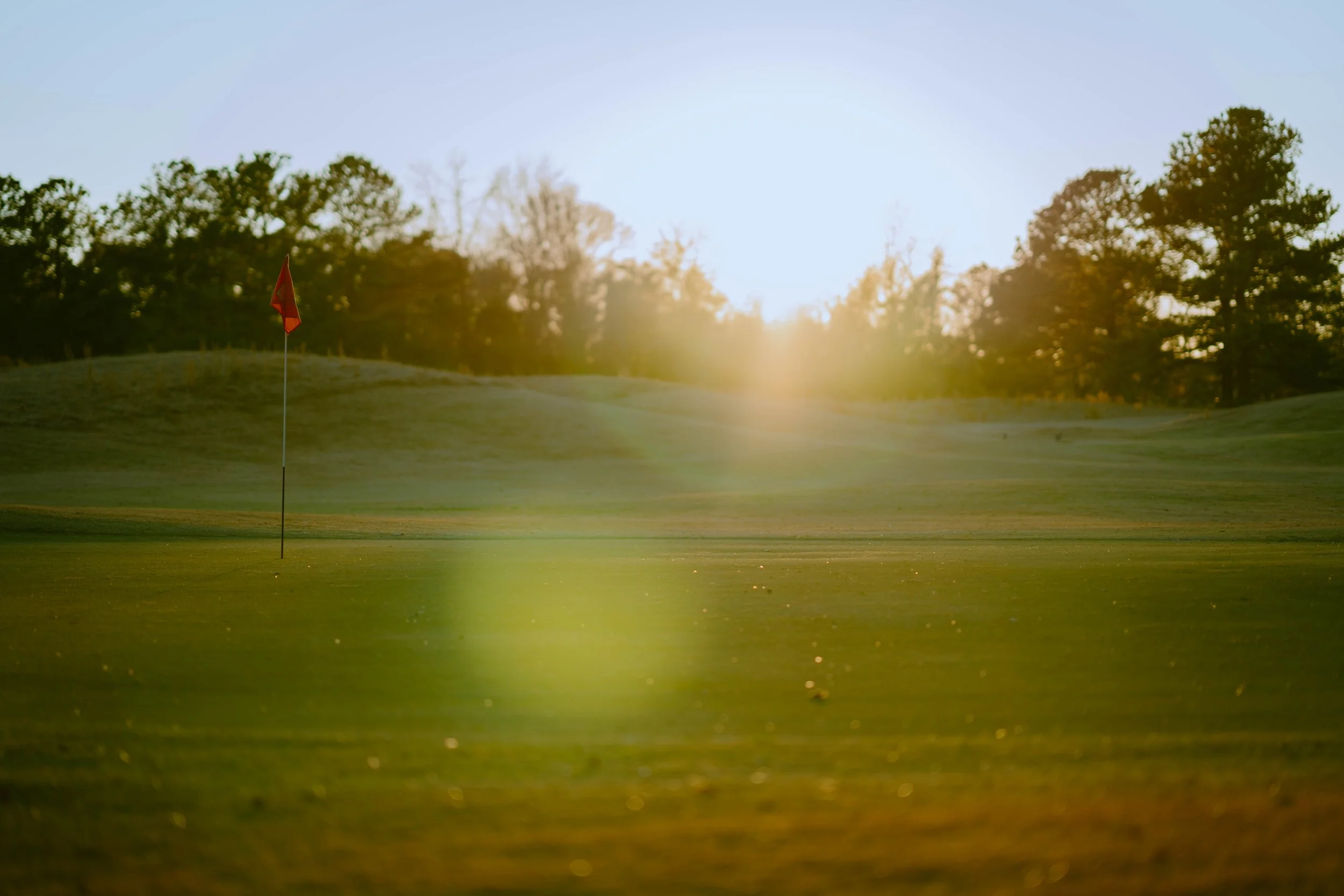 A golf course at sunrise with a red flag on the green and trees in the background.