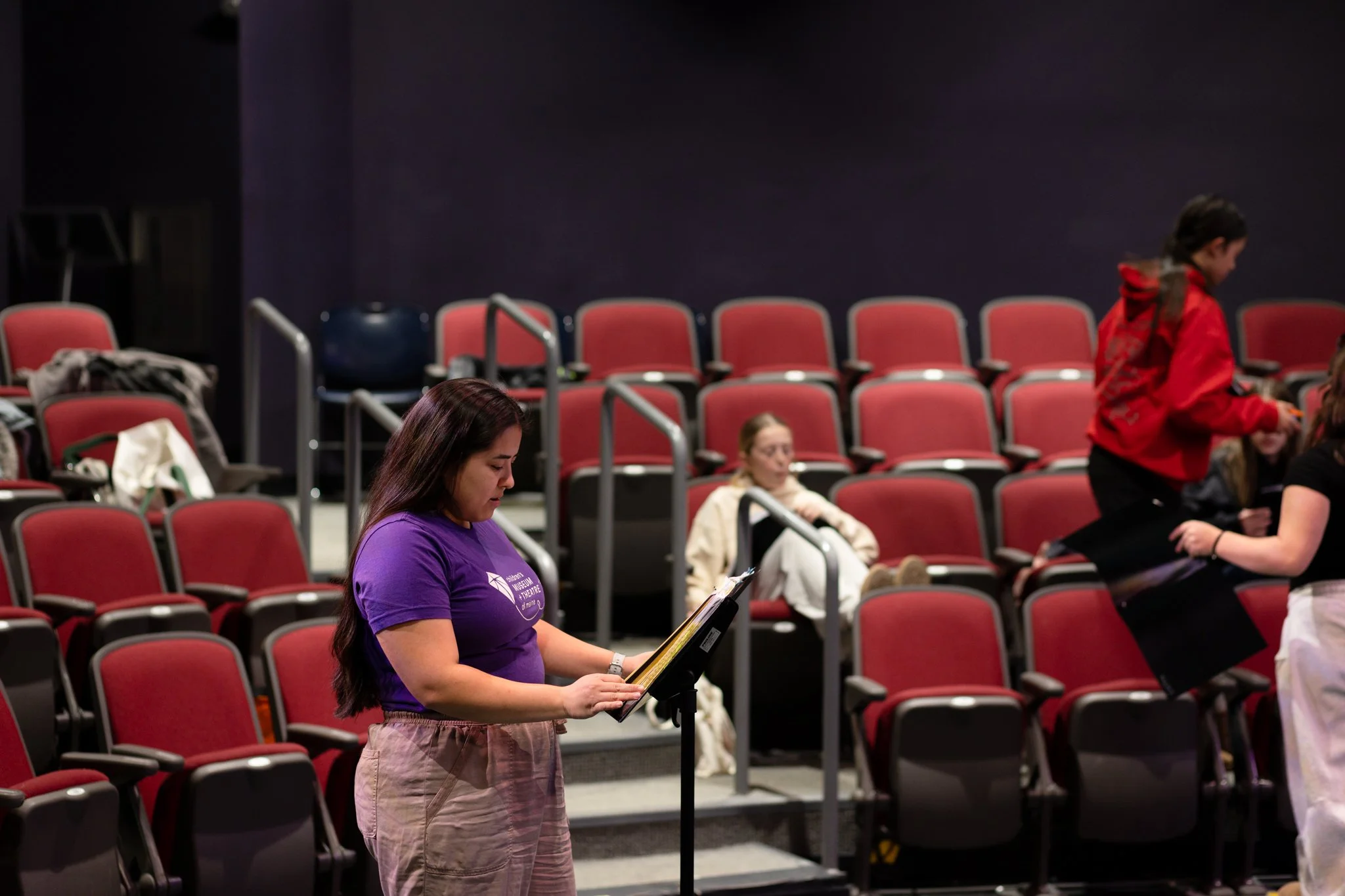 Juliet Tasker stands behind a music stand, directing a scene in SHREK THE MUSICAL: Theatre for Young Audiences Version.