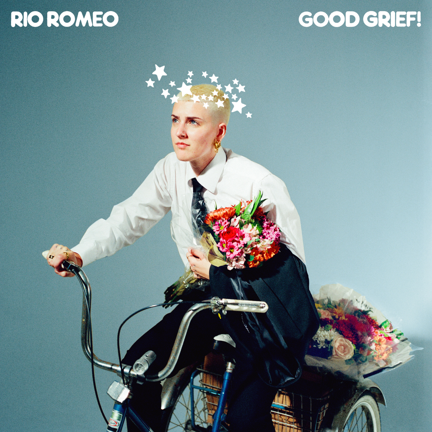 A person with short blonde hair, wearing a white shirt and black tie, is riding a bicycle with a basket filled with colorful flowers. They are holding a bouquet of flowers and have star graphics over their head. The background is plain light blue. The text 'RIO ROMEO' and 'GOOD GRIEF!' are at the top corners of the image.