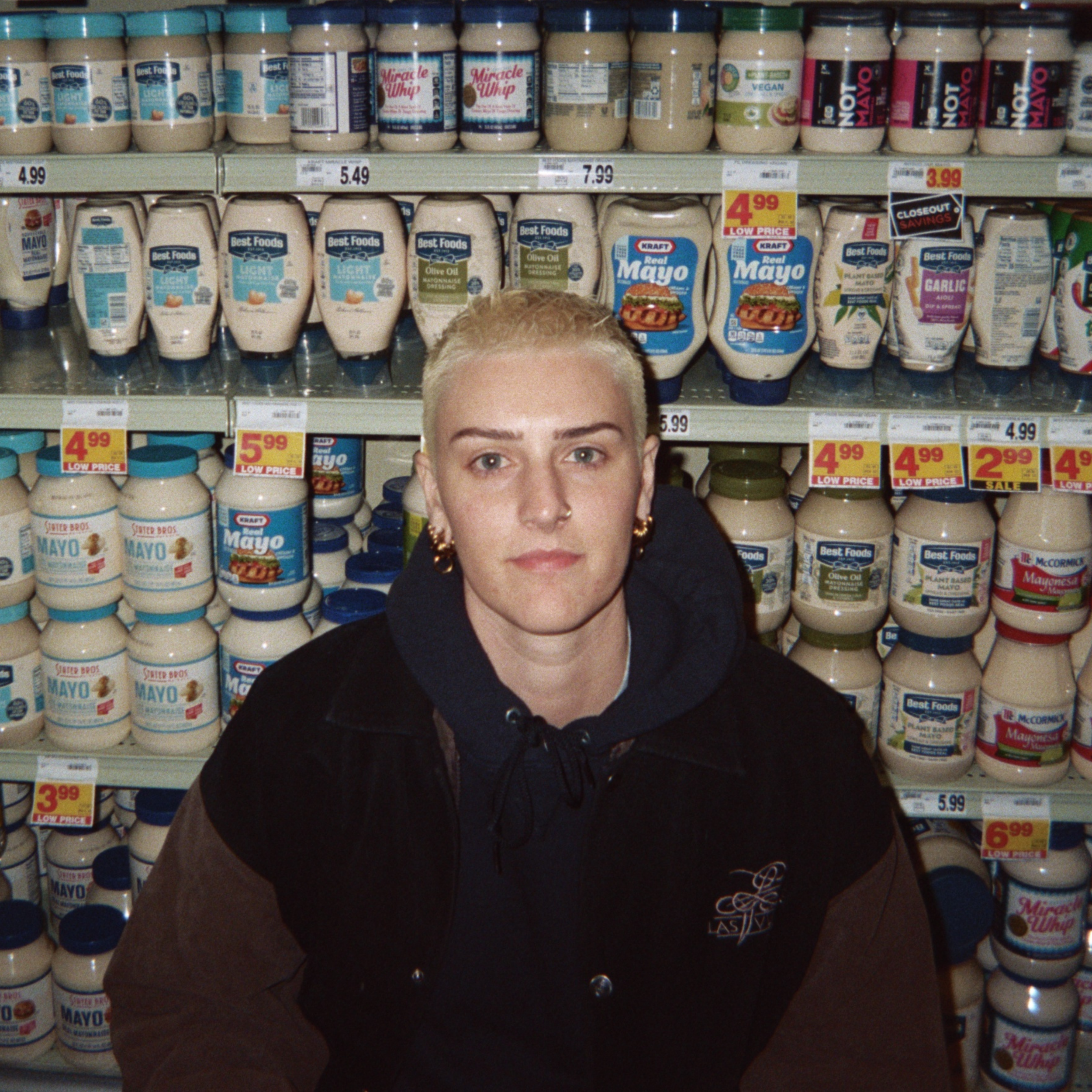 Rio Romeo More than a mother single cover. Rio Romeo, blonde buzzcut butch with gold jewelry and a black hoodie, is standing in front of a grocery shelf full of mayo bottles and jars.