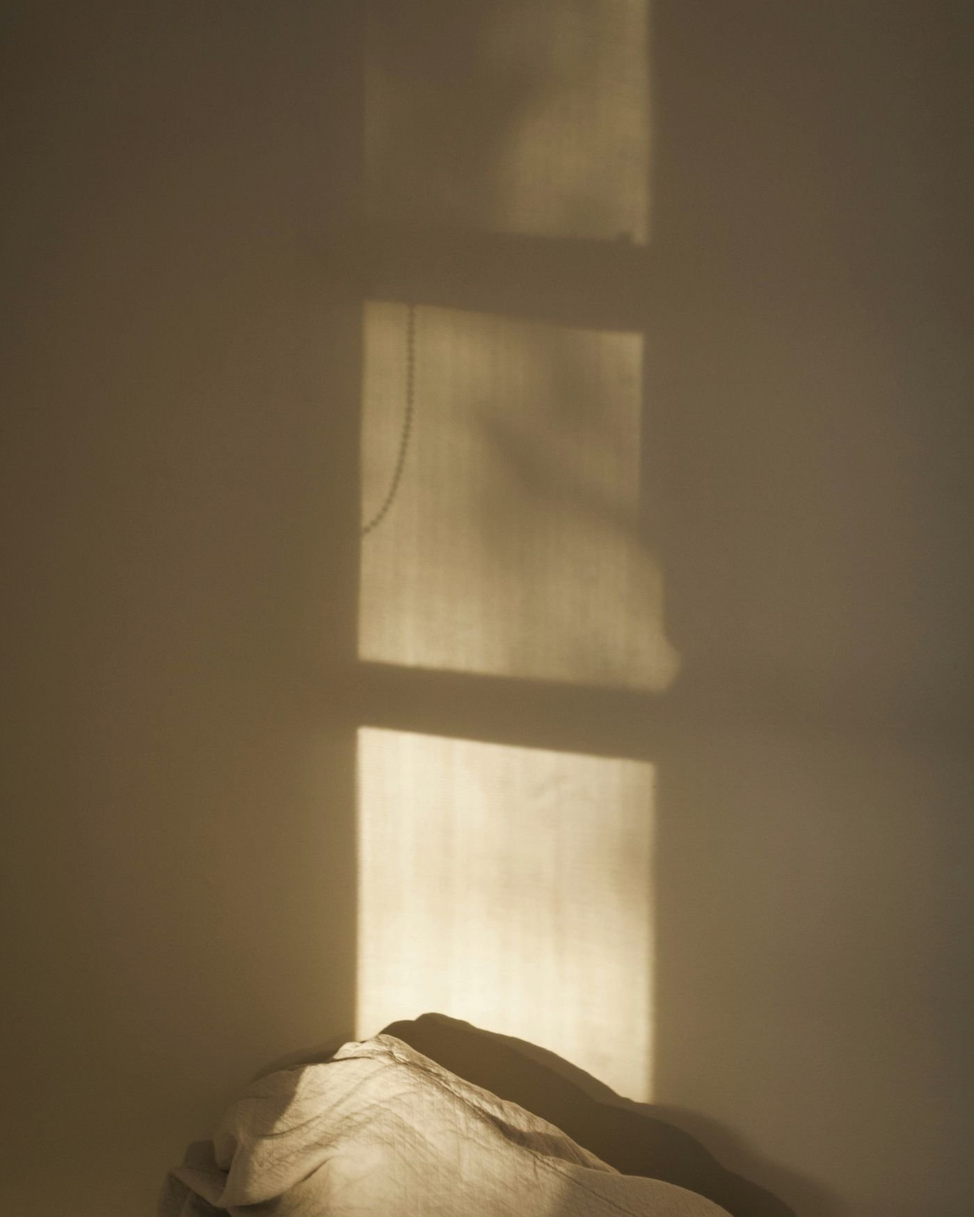 Soft warm lights falls on a wall above a linen pillow on an unmade bed.