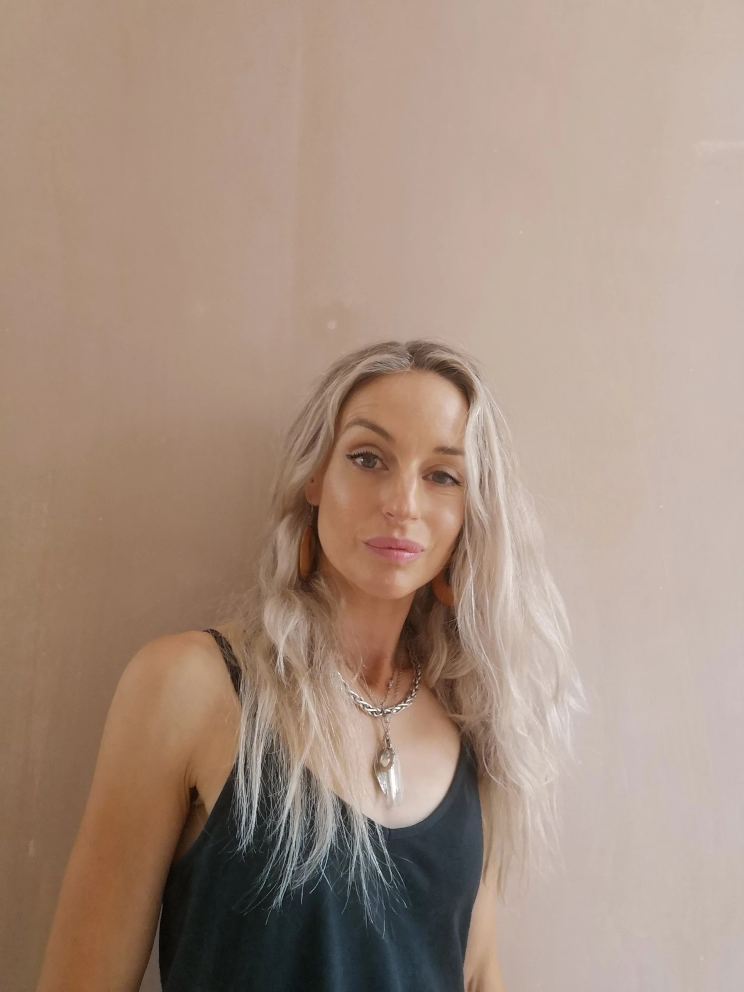 A woman with long, wavy blonde hair wearing a black sleeveless top, layered necklaces, and orange earrings standing against a beige wall.