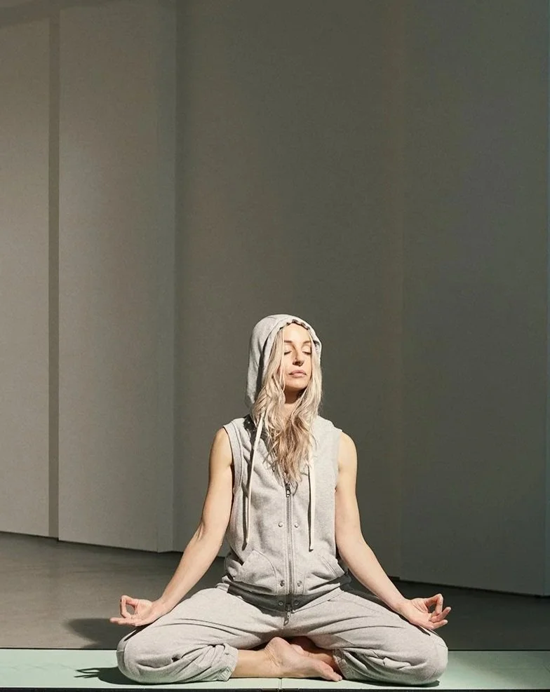 A woman with long blonde hair, wearing a gray sleeveless hoodie and sweatpants, sitting cross-legged in a yoga pose on a green mat with her eyes closed in a minimalist room.