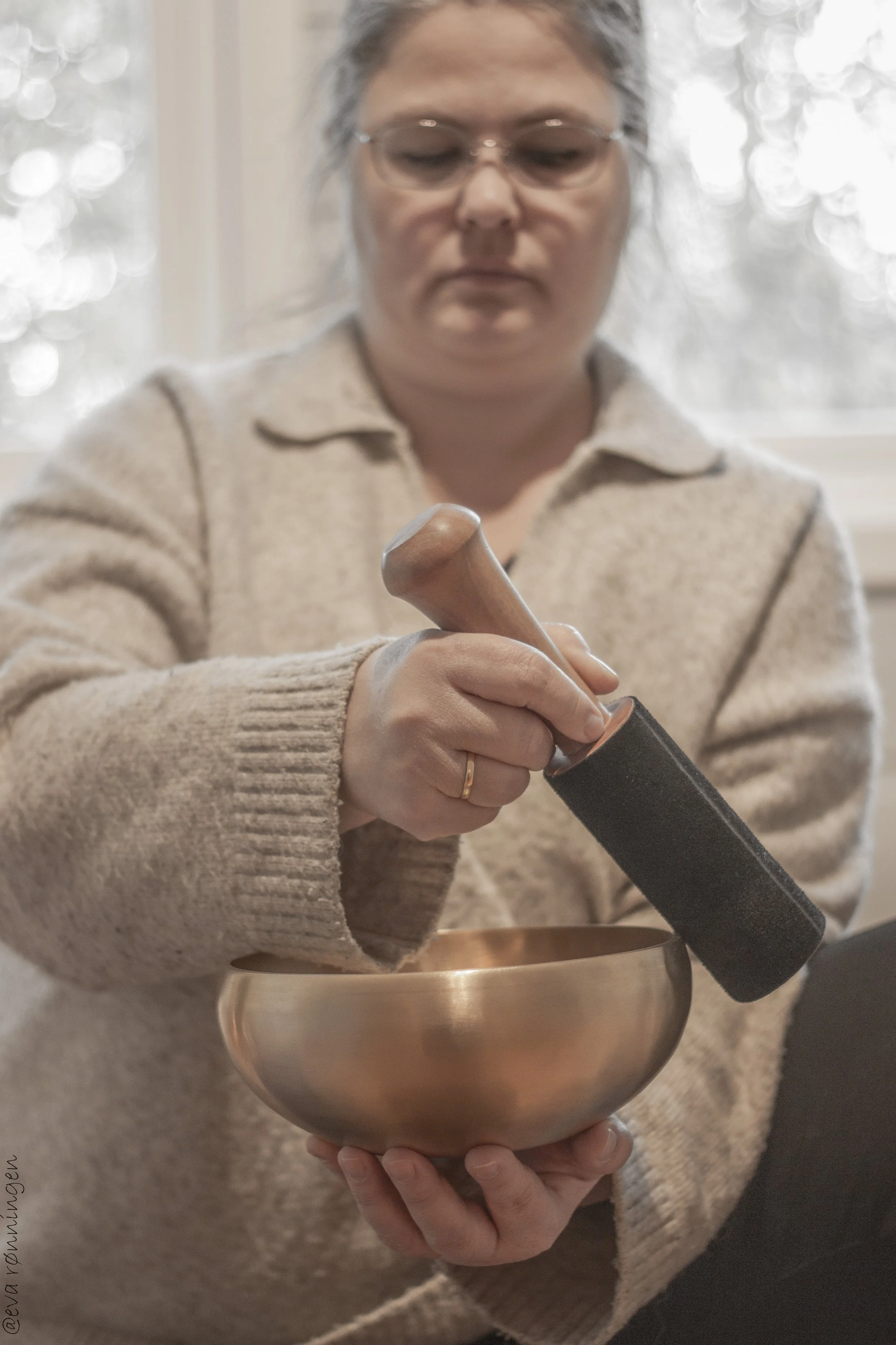 Nina sitter og holder en klangskål. Øynene er lukket og uttrykket er konsentrert og avslappet. Lyset er mykt, med et vindu i bakgrunnen som gir en rolig og dempet stemning.