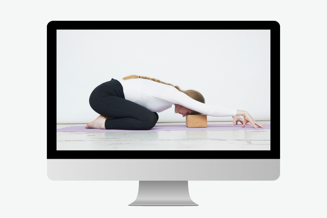 The images shows me practicing on a purple mat, shown on a computer. I am wearing black trousers and a white top. My head is resting on a yoga block.