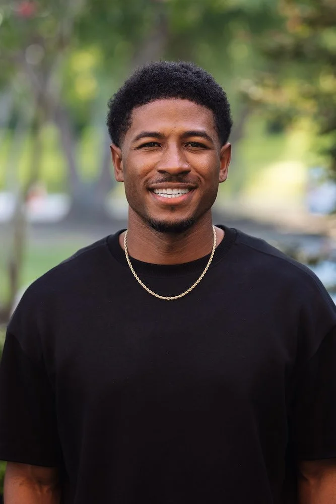 A smiling young man with short curly hair, wearing a black t-shirt and a gold chain, standing outdoors in a park with trees and blurred background.