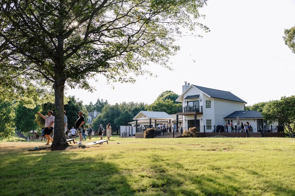 People gathered outdoors near a white house with a deck, trees, and grassy area in a residential neighborhood.
