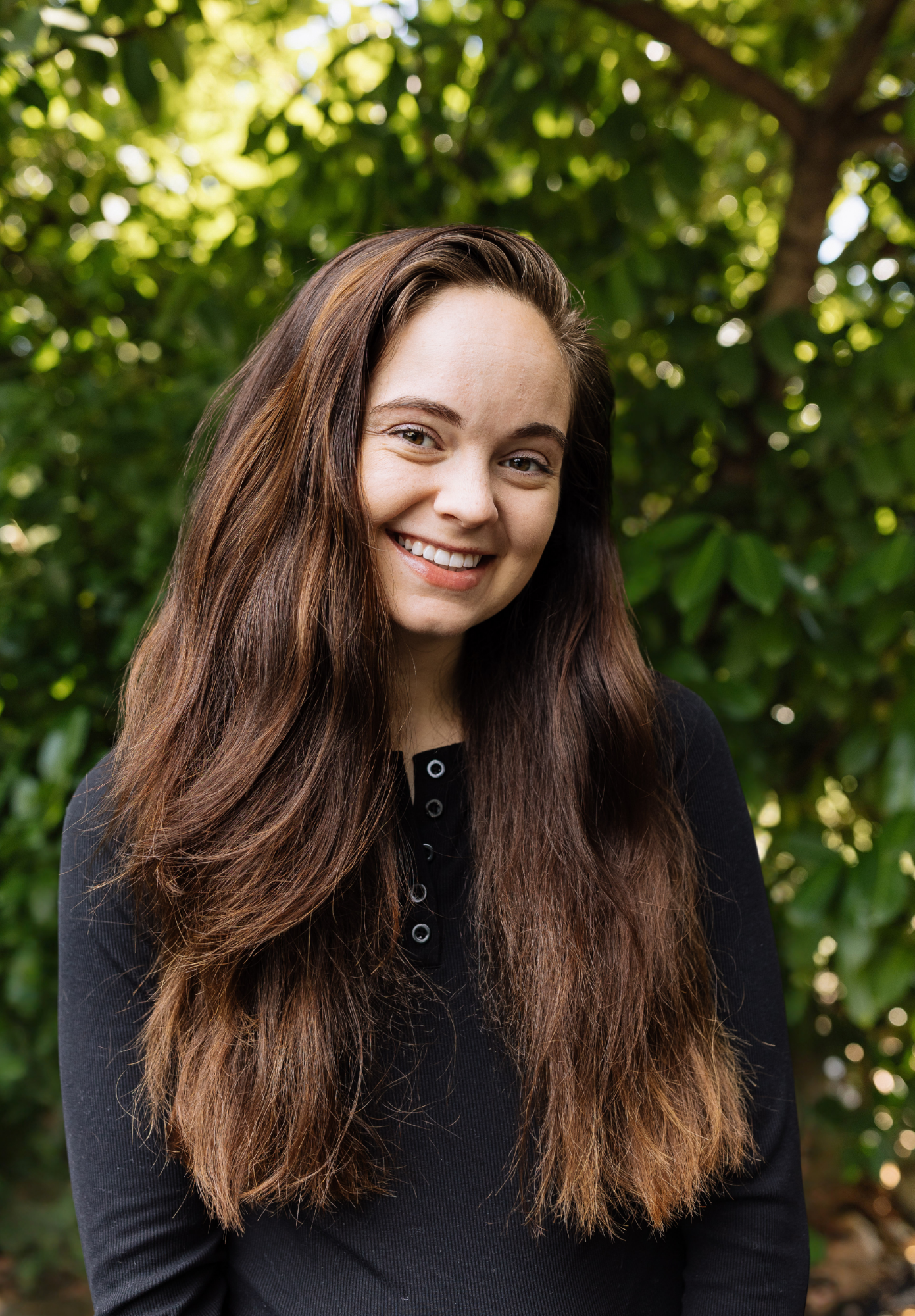 A young woman with long, wavy auburn hair smiling and looking over her shoulder outdoors, wearing a black off-the-shoulder top.