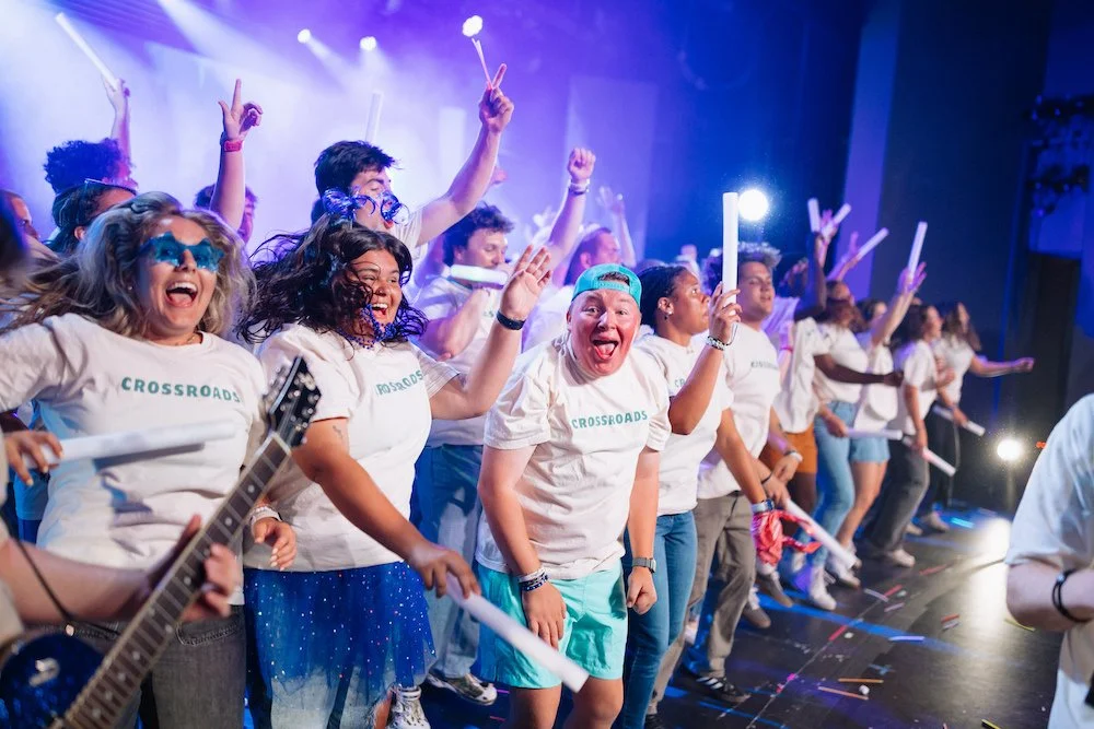 A group of joyful people on stage wearing matching white T-shirts with the word "CROSSROADS" printed on them, celebrating with raised hands and paper glow sticks, with colorful stage lighting.