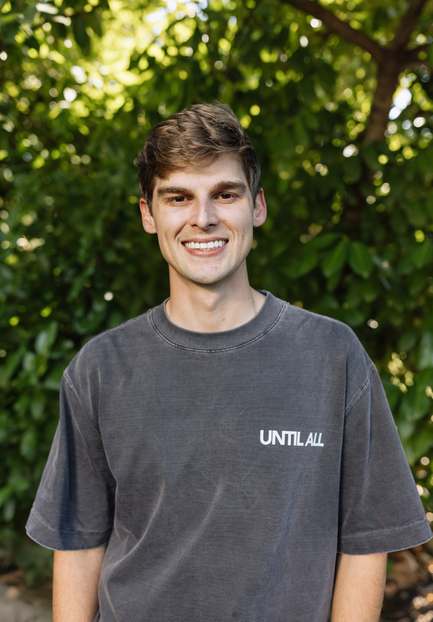 A young man with short brown hair and a big smile, wearing a dark gray T-shirt with the words 'UNTIL ALL' printed on it, standing outdoors with green trees in the background.