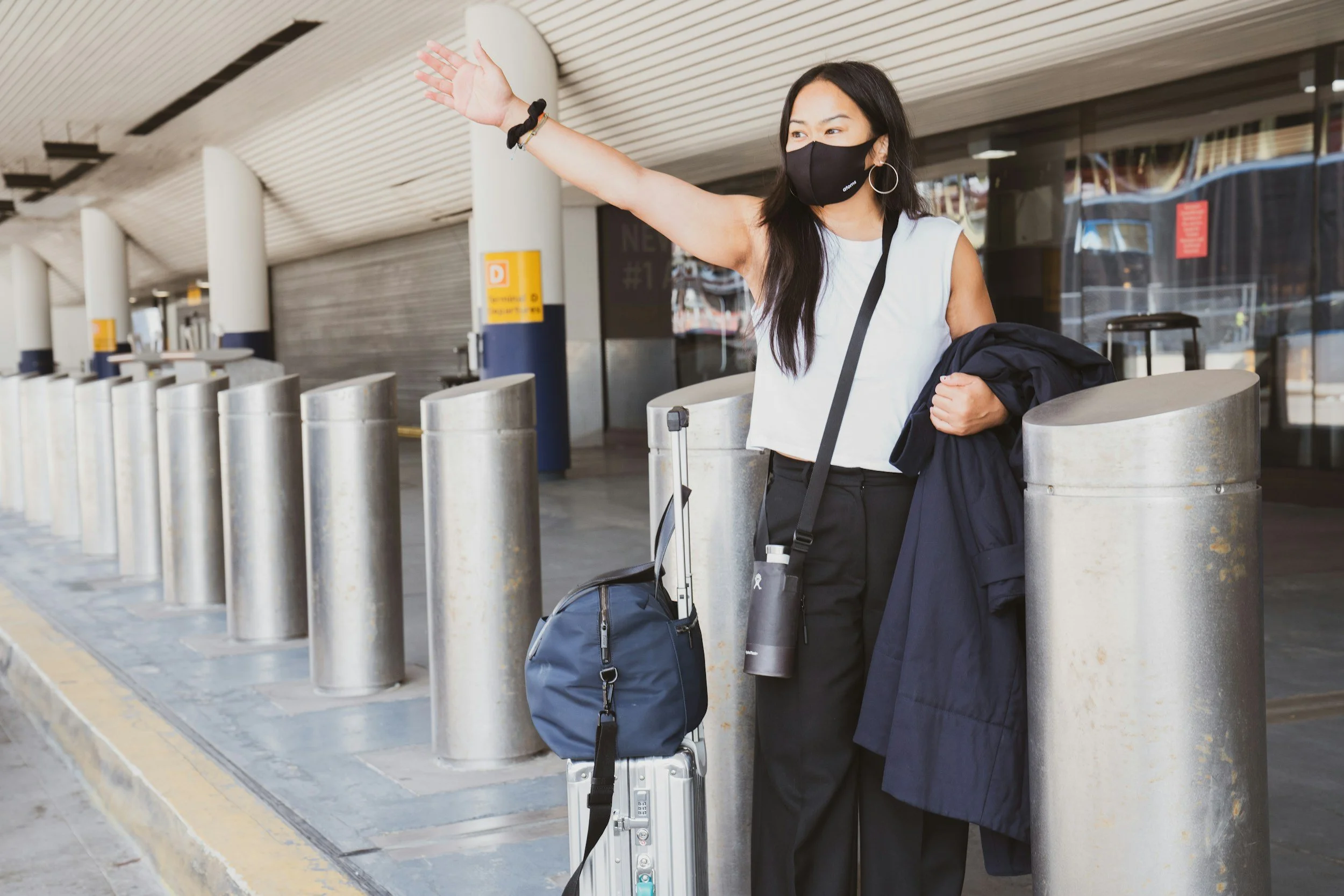 A woman wearing a black face mask, black pants, and a white sleeveless top, standing at an airport terminal, waving with her right hand, holding a blue bag and gray suitcase, with a black jacket and a black water bottle in her left arm, near airport security barriers.