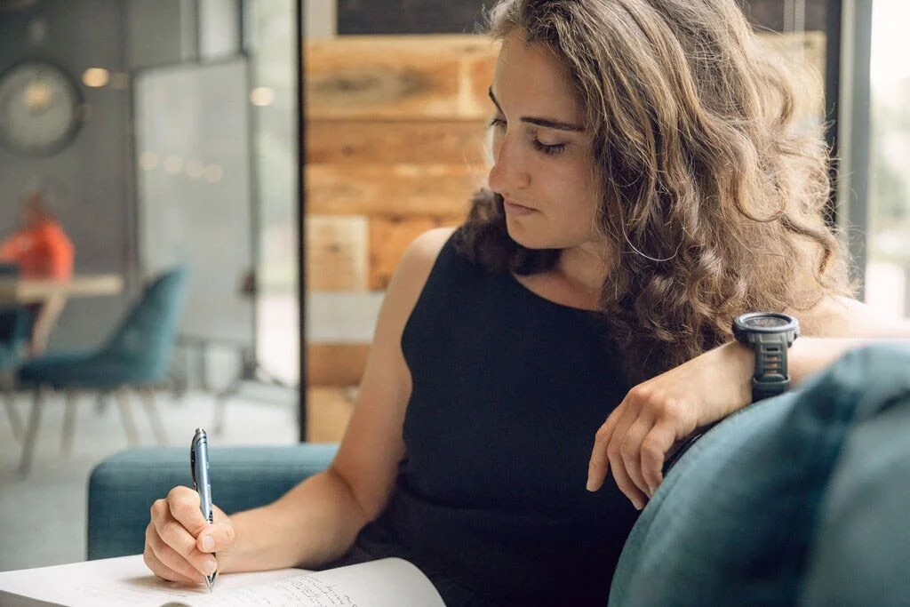 Young woman with curly hair writing in a notebook with a pen, sitting on a teal sofa in a modern, cozy room.