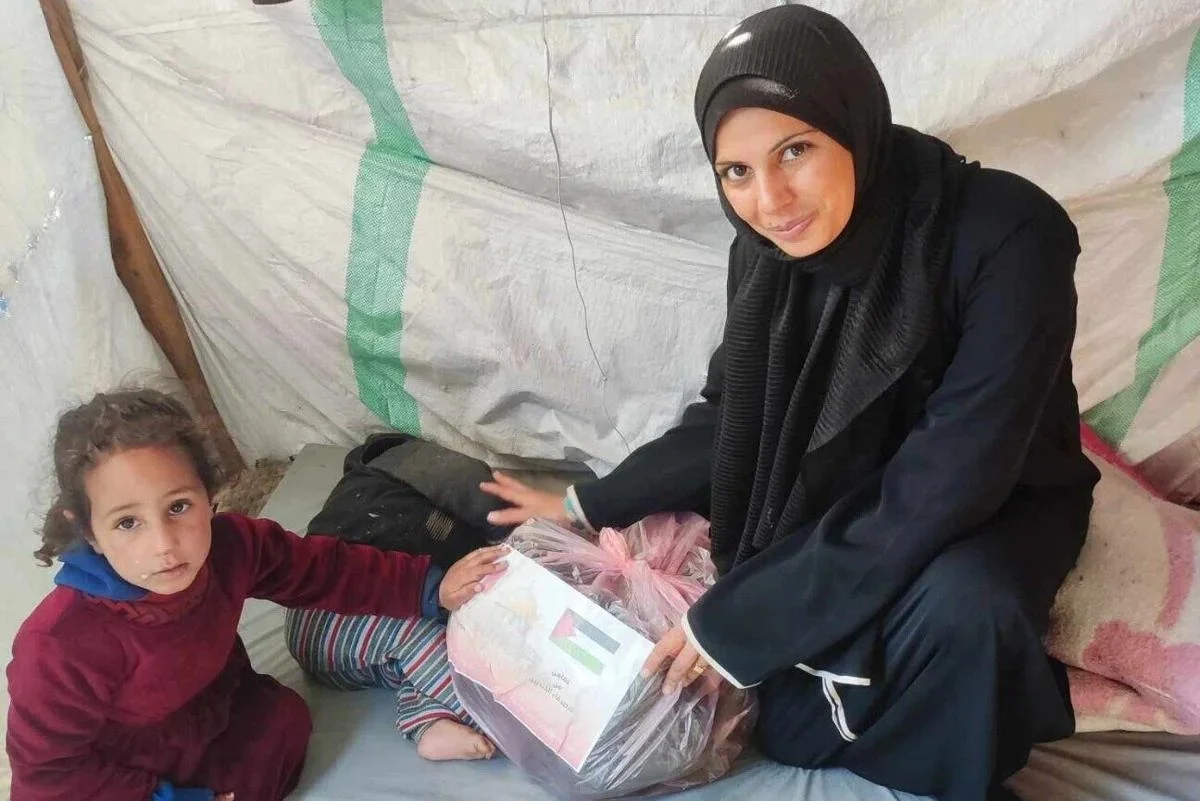 Woman and child in a tent with a packaged food item, displaying a flag and writing in Arabic.