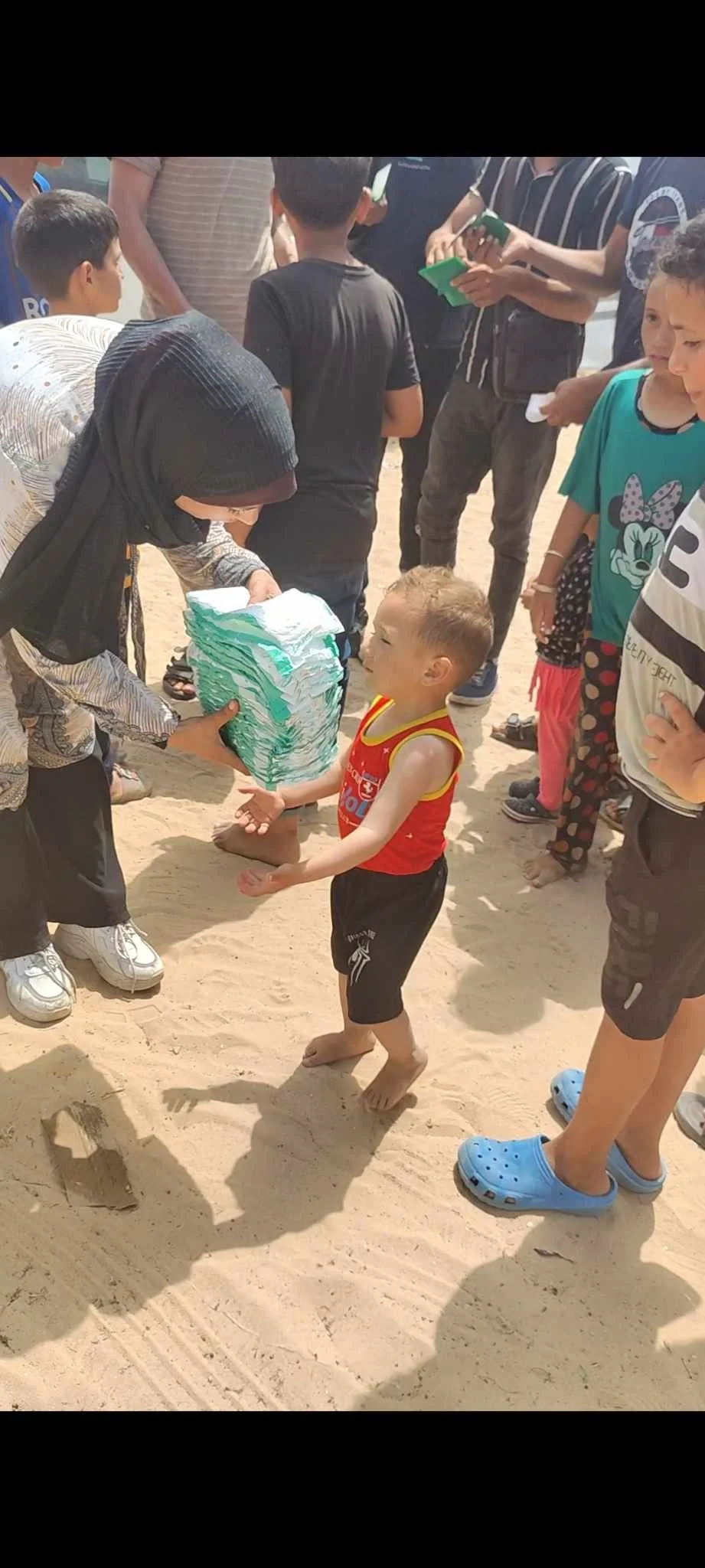 A woman handing stacks of diapers to a young boy on sand, surrounded by other children and adults.