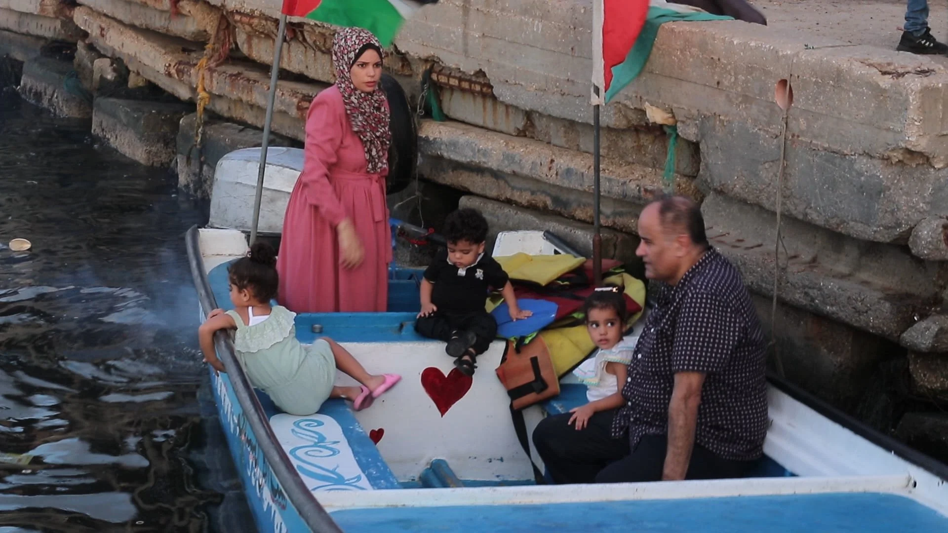 A family with children sitting in a small boat near a concrete dock, with flags in the background.