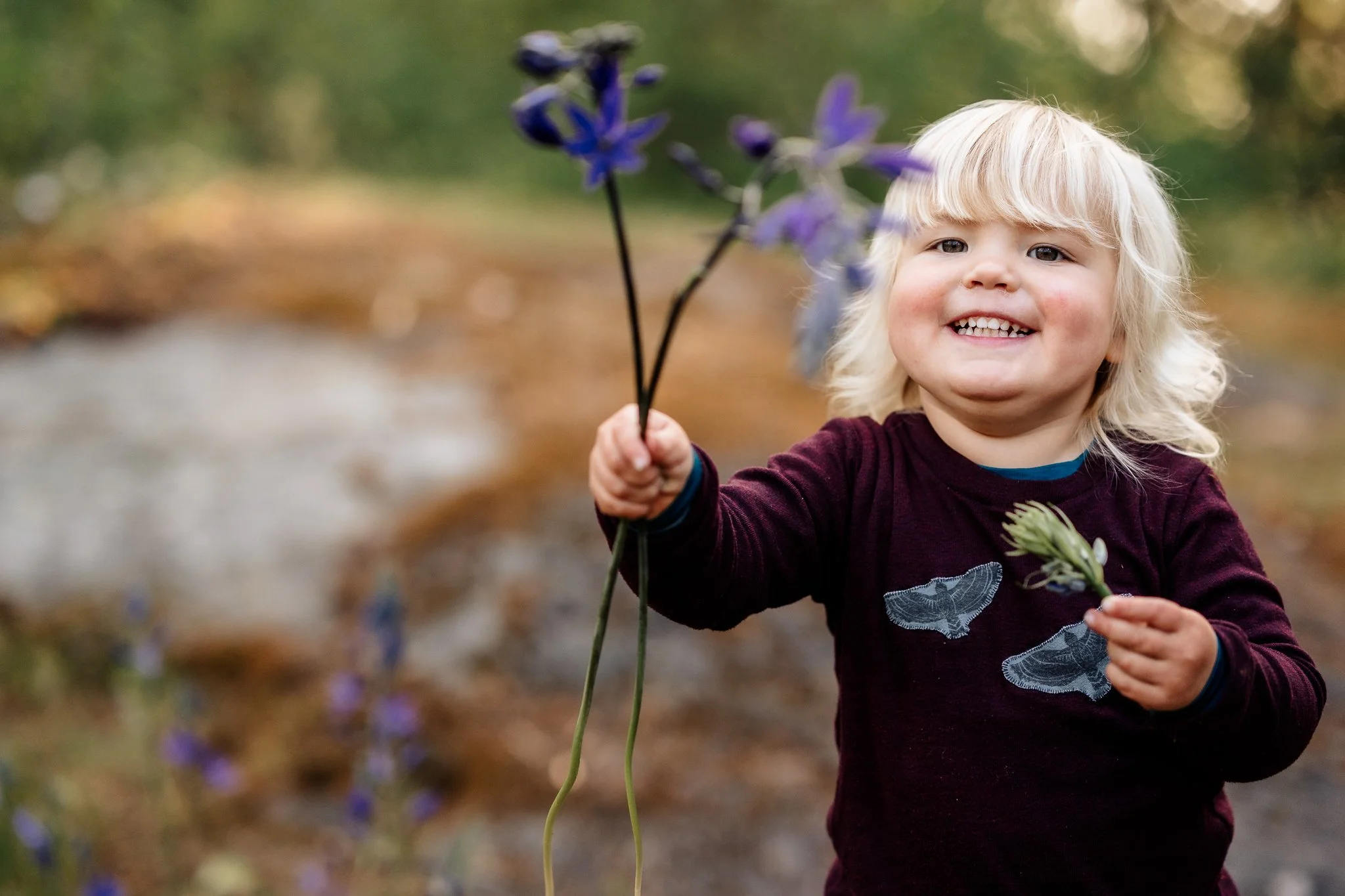 A smiling toddler with blonde hair holding purple flowers outdoors.